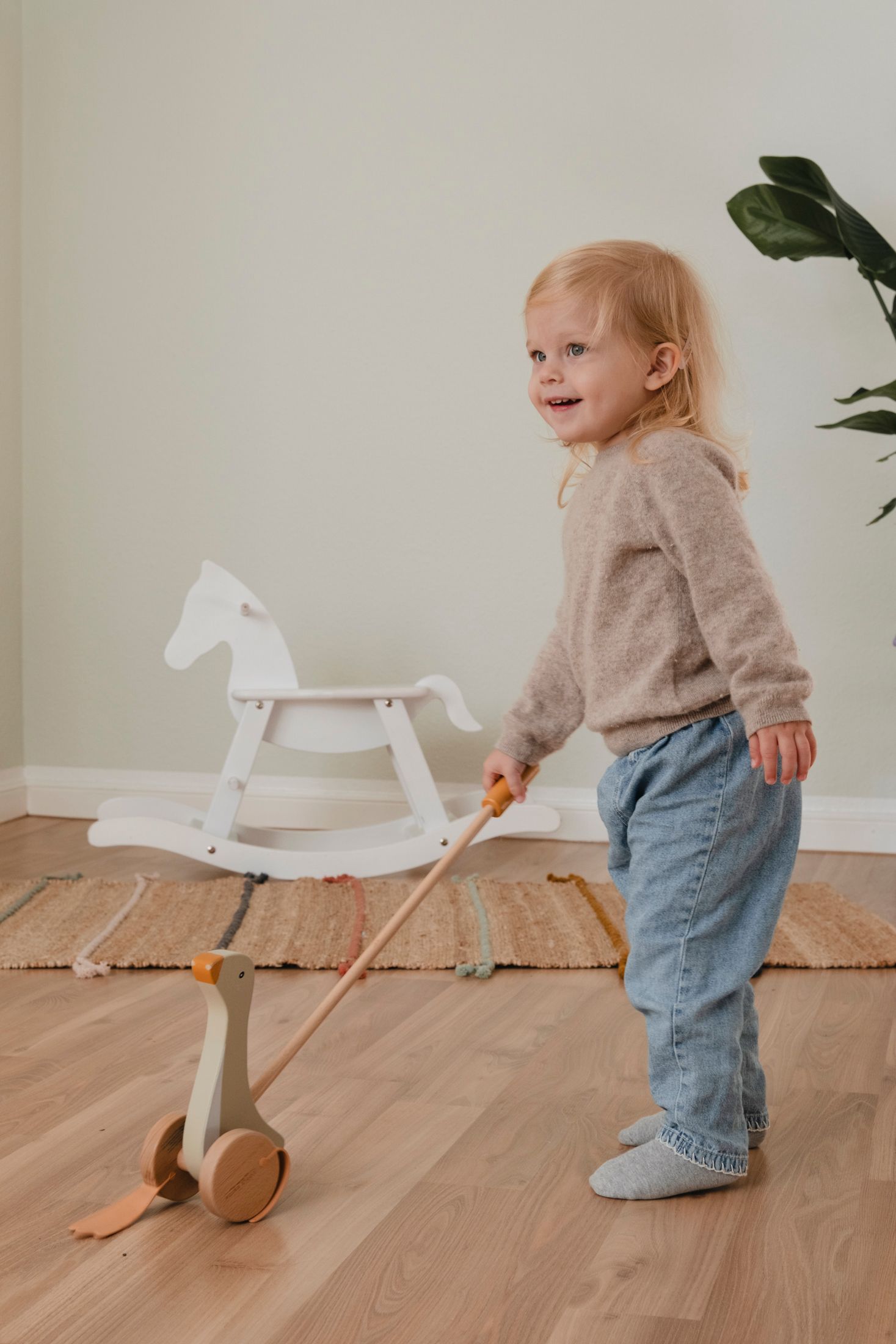 Smiling blonde toddler girl pushing wooden duck pull toy with rocking horse in background.