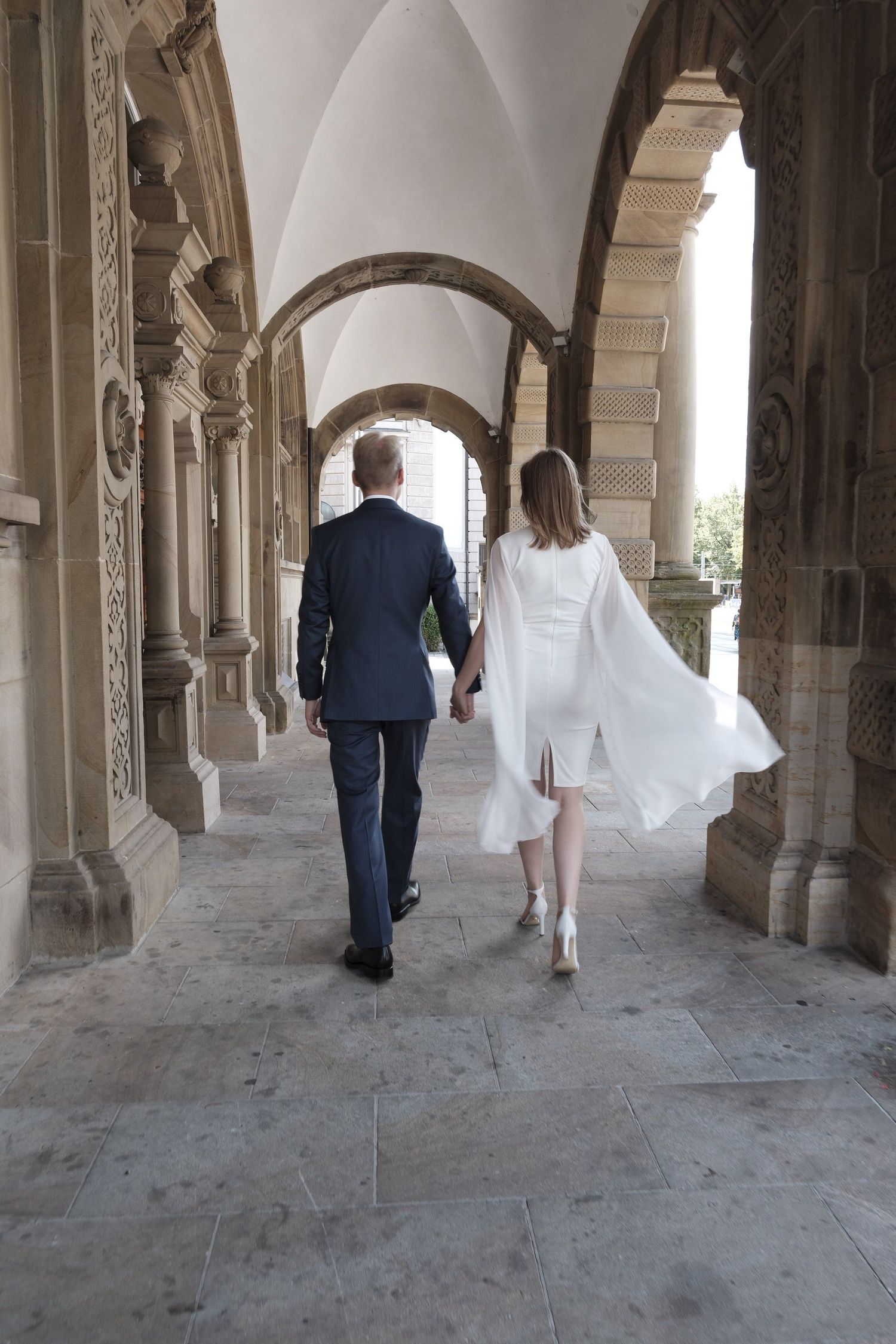 Bride and groom walking hand in hand through elegant stone archway corridor.