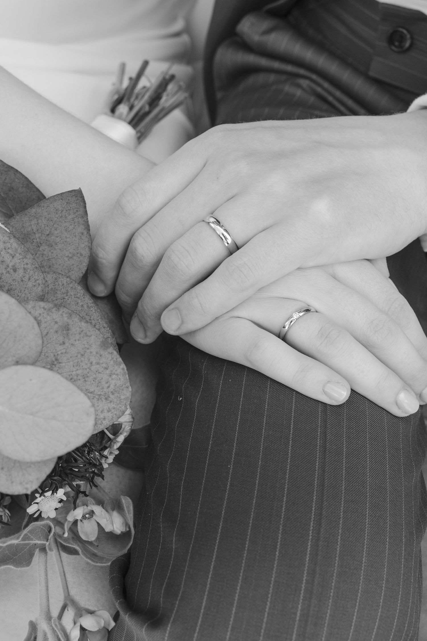 Black and white close-up of newlyweds hands displaying matching silver wedding rings beside a bouquet.