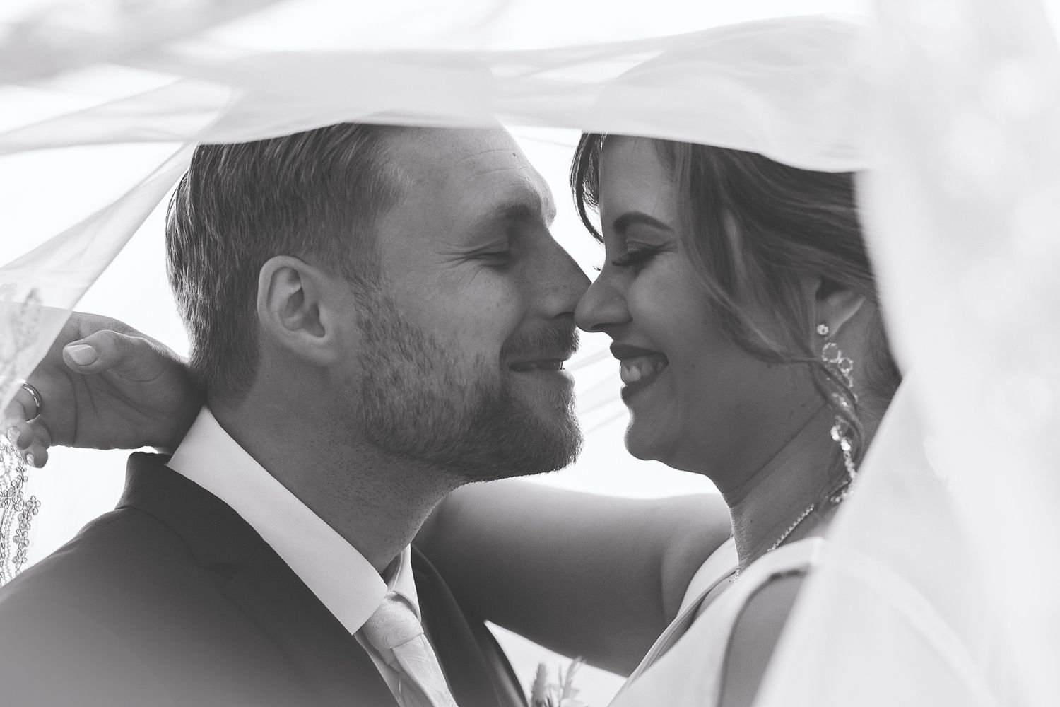 Black and white romantic photo of bride and groom almost kissing under wedding veil.