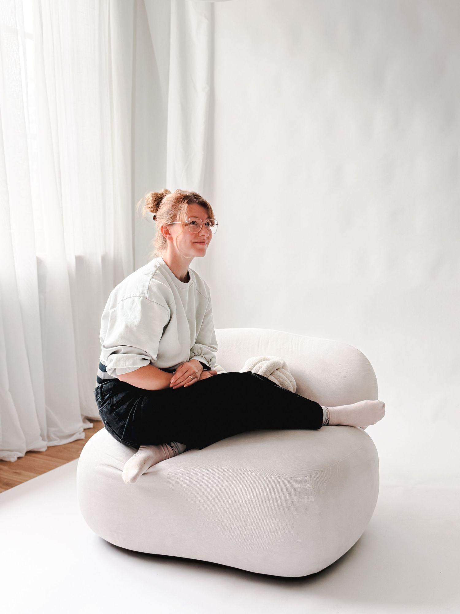 Young woman with glasses sitting cross-legged on a modern white boucle armchair indoors.