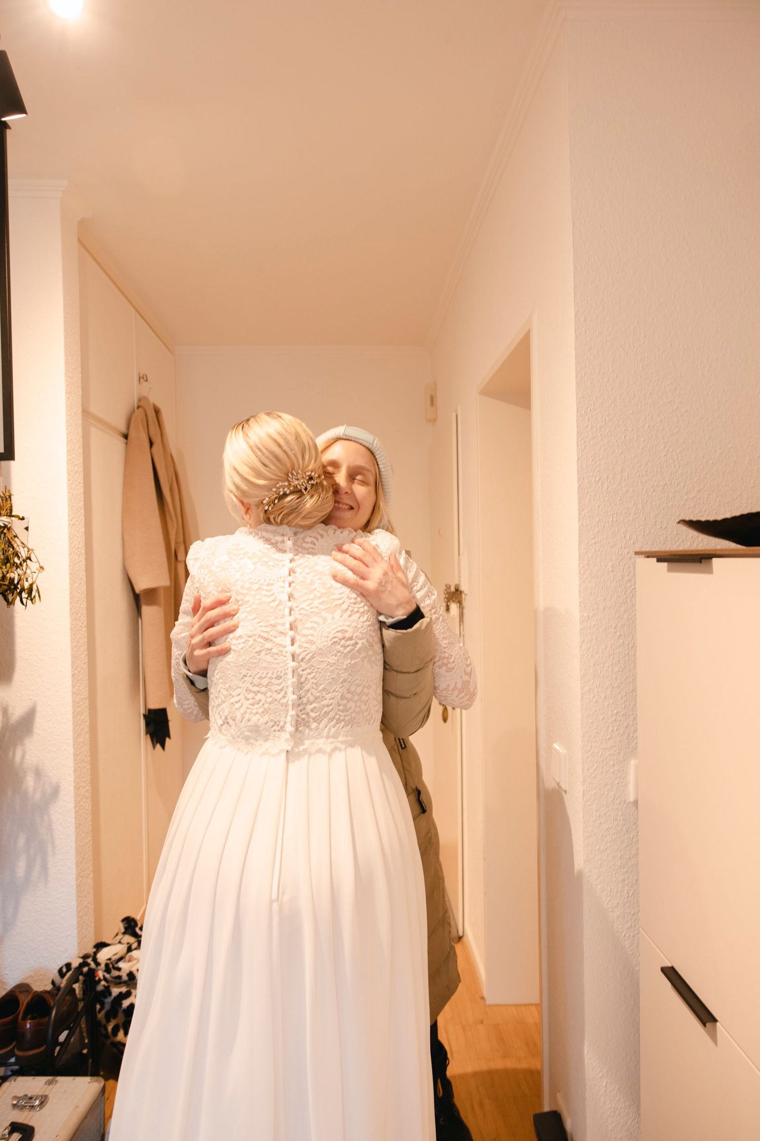 Bride in white lace wedding dress shares warm embrace with guest in hallway.