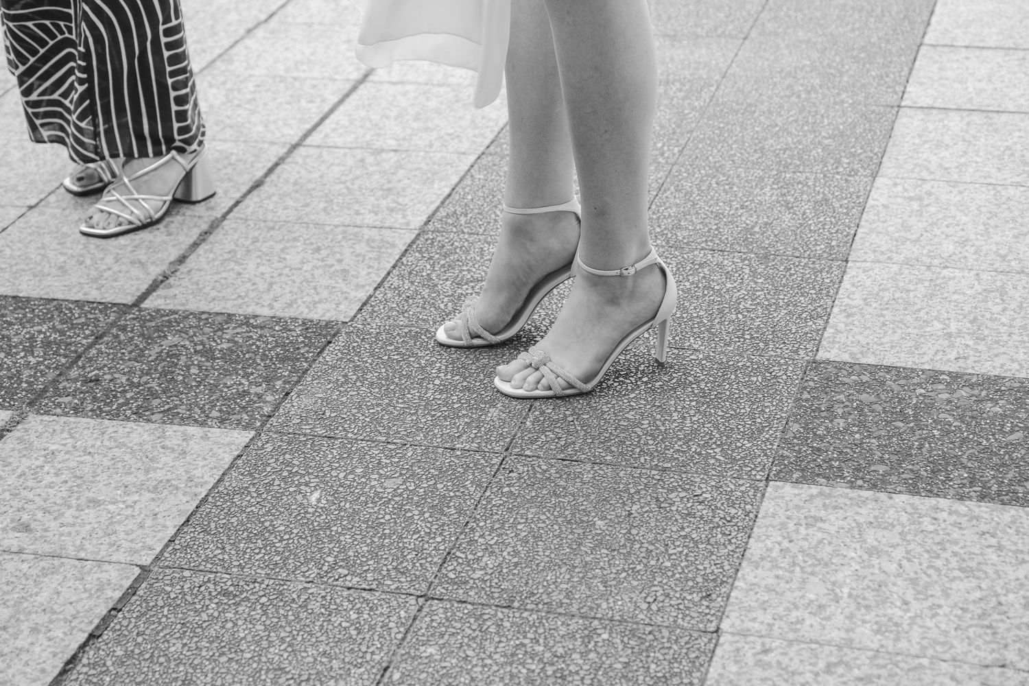 Black and white close-up of a bride wearing elegant strappy heels on a tiled surface.