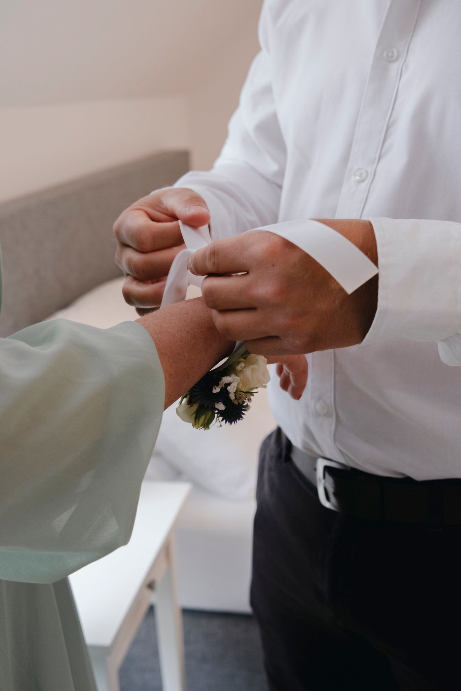 Close-up of hands attaching a floral corsage with white ribbon to a mans shirt cuff.