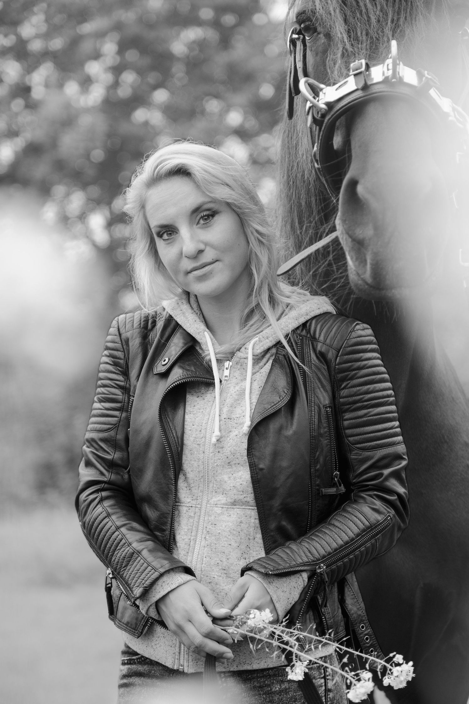 Black and white photo of a blonde woman in a leather jacket holding flowers beside a horse outdoors.