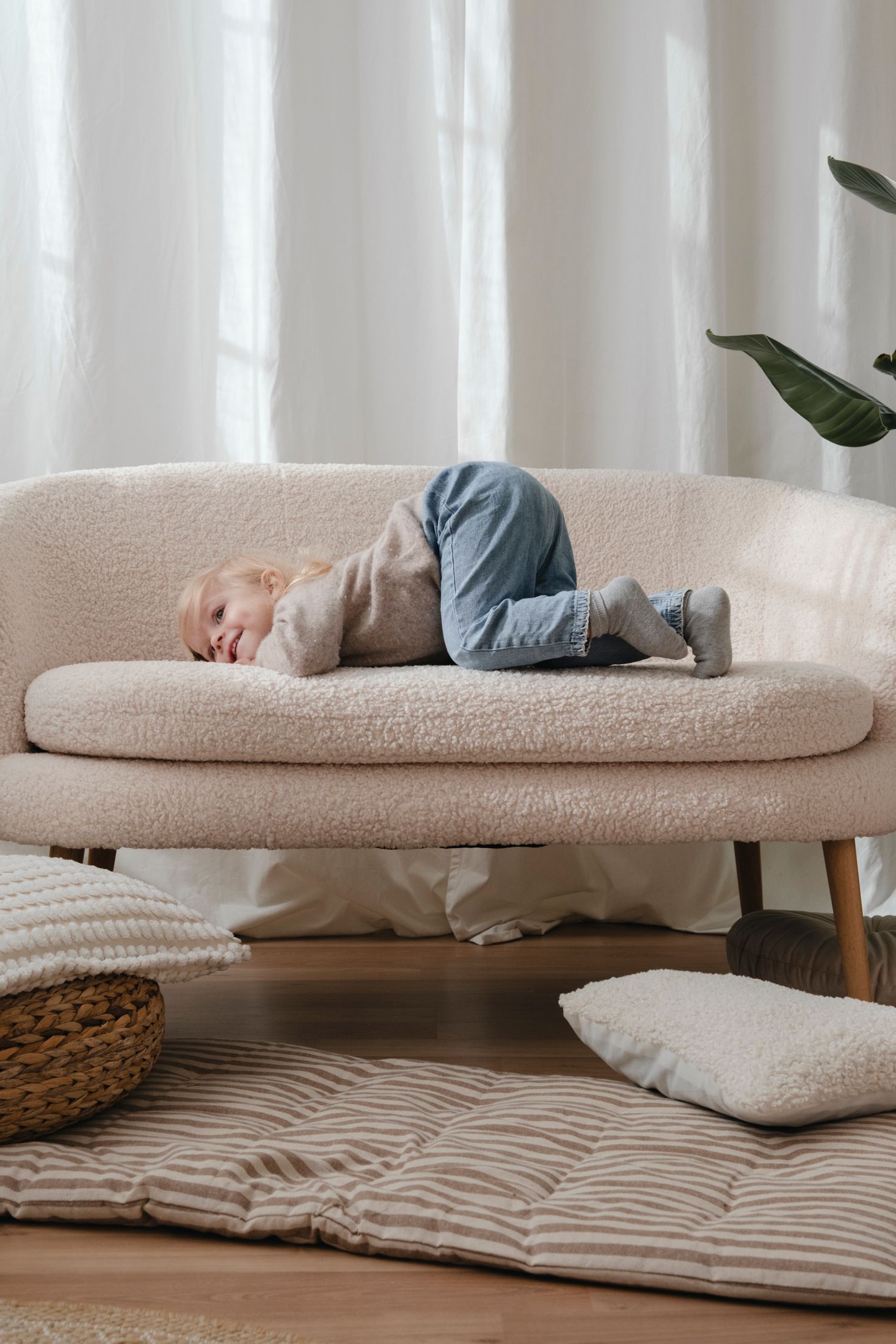 Blonde toddler lying curled up on cream boucle sofa in bright natural light living room.