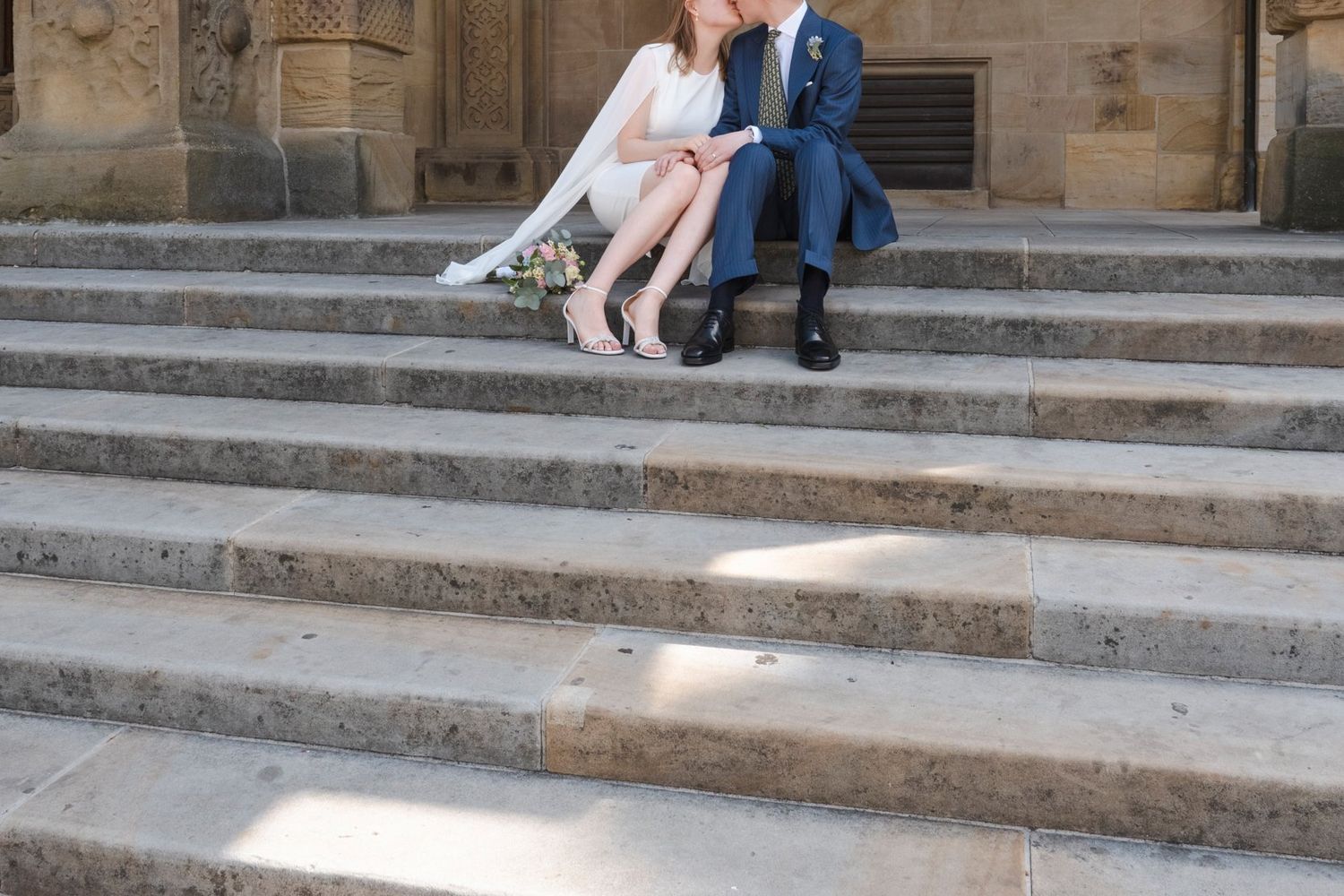 Bride and groom sitting closely together on stone steps of a historic building holding a floral bouquet.