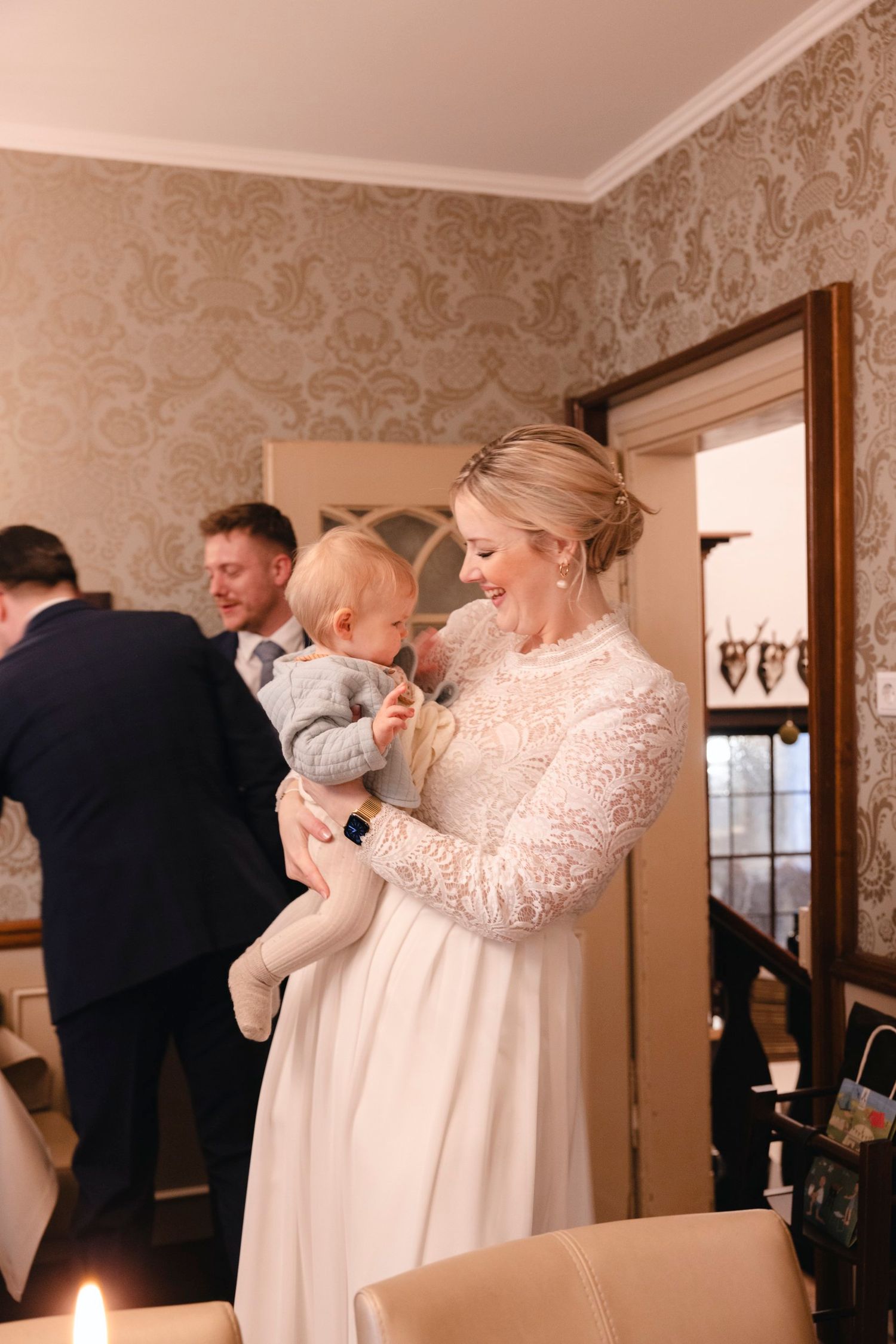 Smiling bride in lace long sleeve wedding dress tenderly holding baby at intimate winter wedding reception.