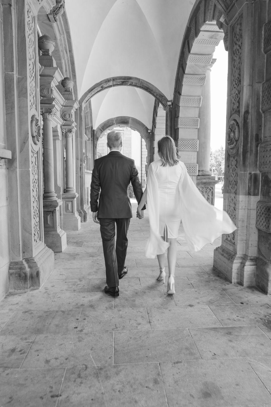 A black and white photo of a bride and groom walking through an ornate stone archway.