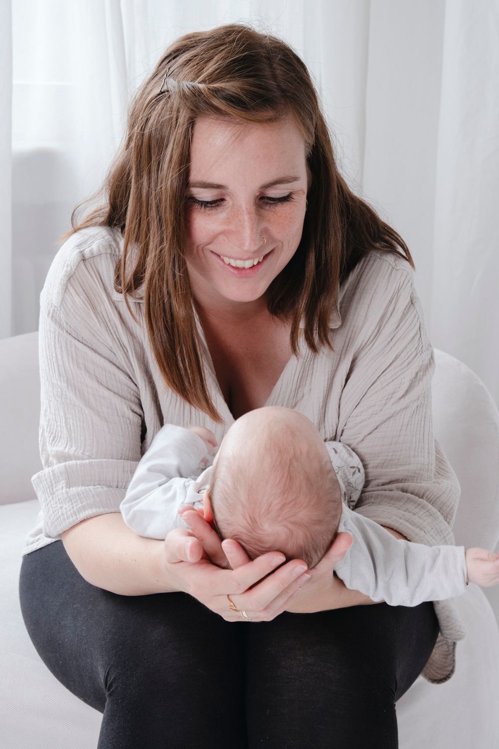 Smiling young mother holding her newborn baby on a white chair in a bright room.