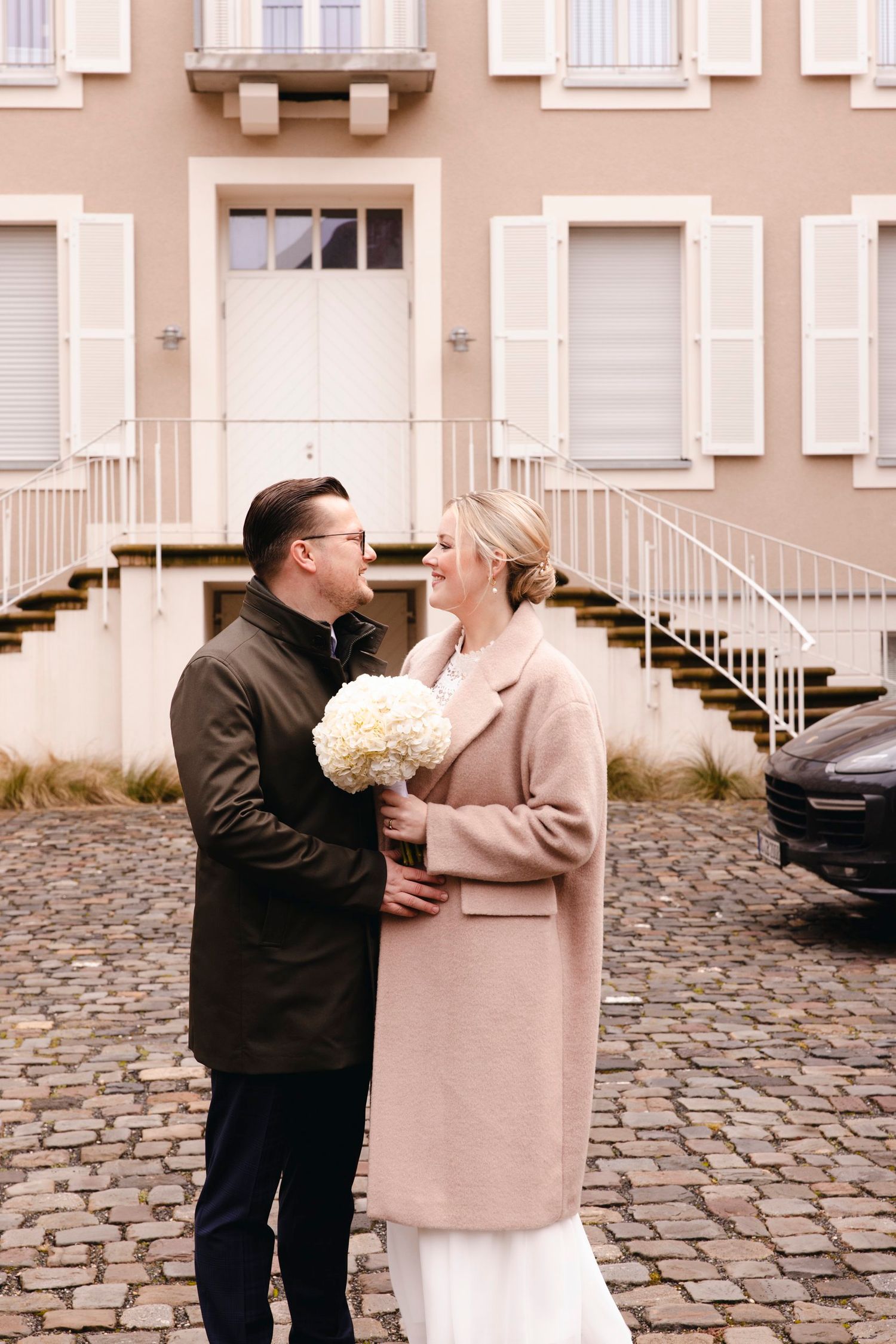 Bride and groom share a romantic moment outdoors on cobblestones, bride holding white hydrangea bouquet wearing pink coat.
