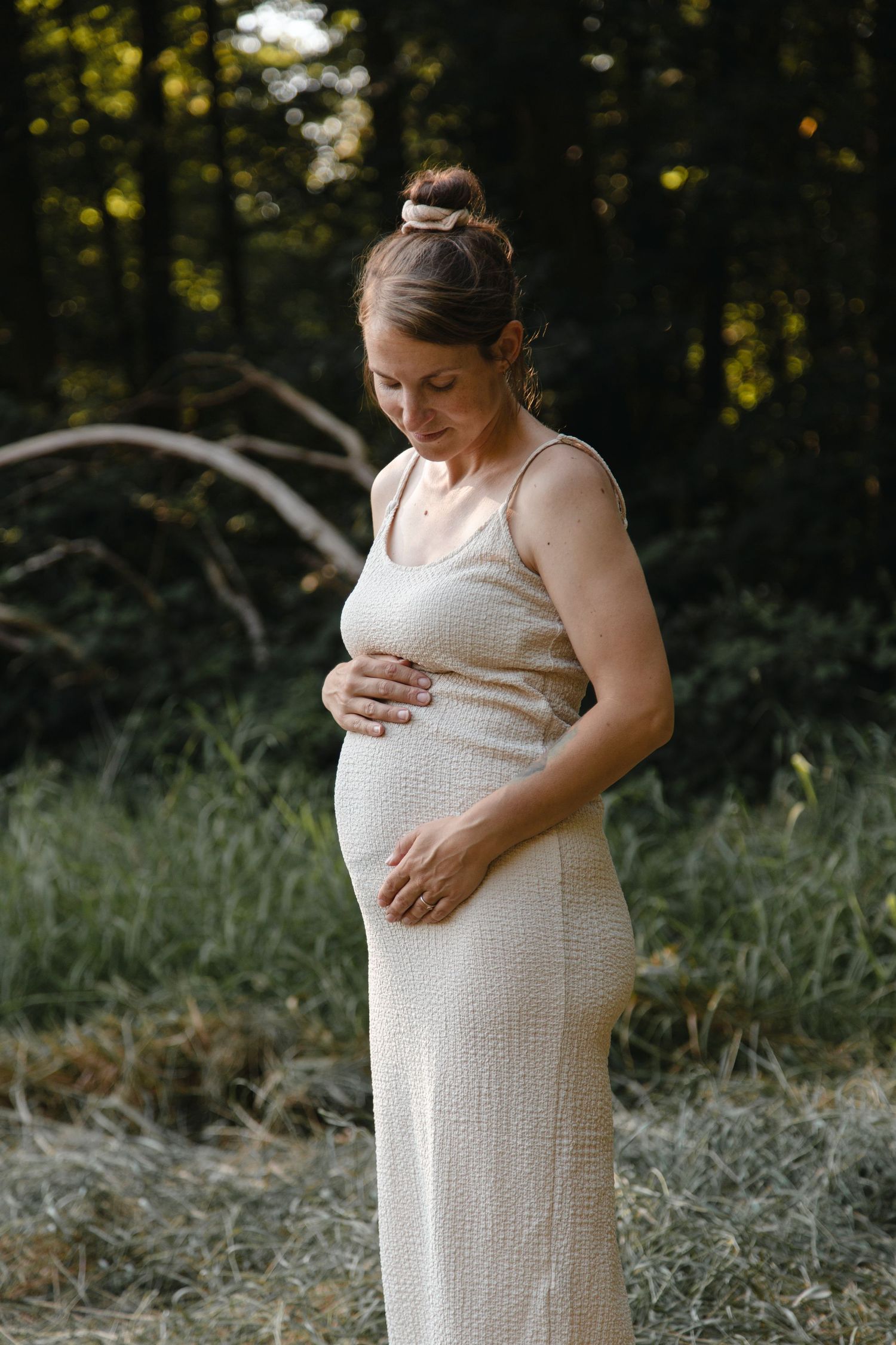 Pregnant woman in cream knit dress cradling baby bump while standing in forest clearing.