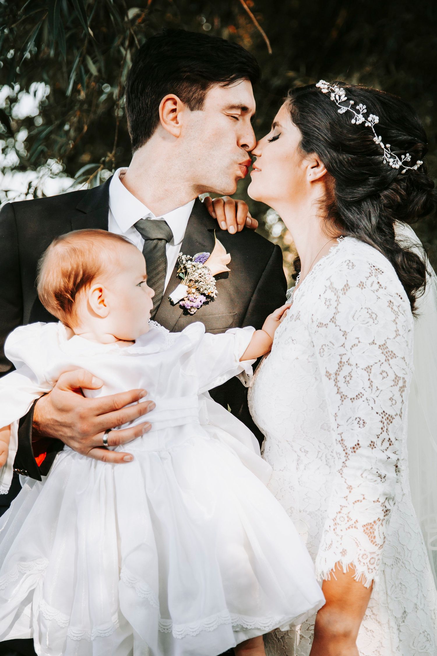 Wedding couple kissing while holding their baby dressed in white during outdoor ceremony.