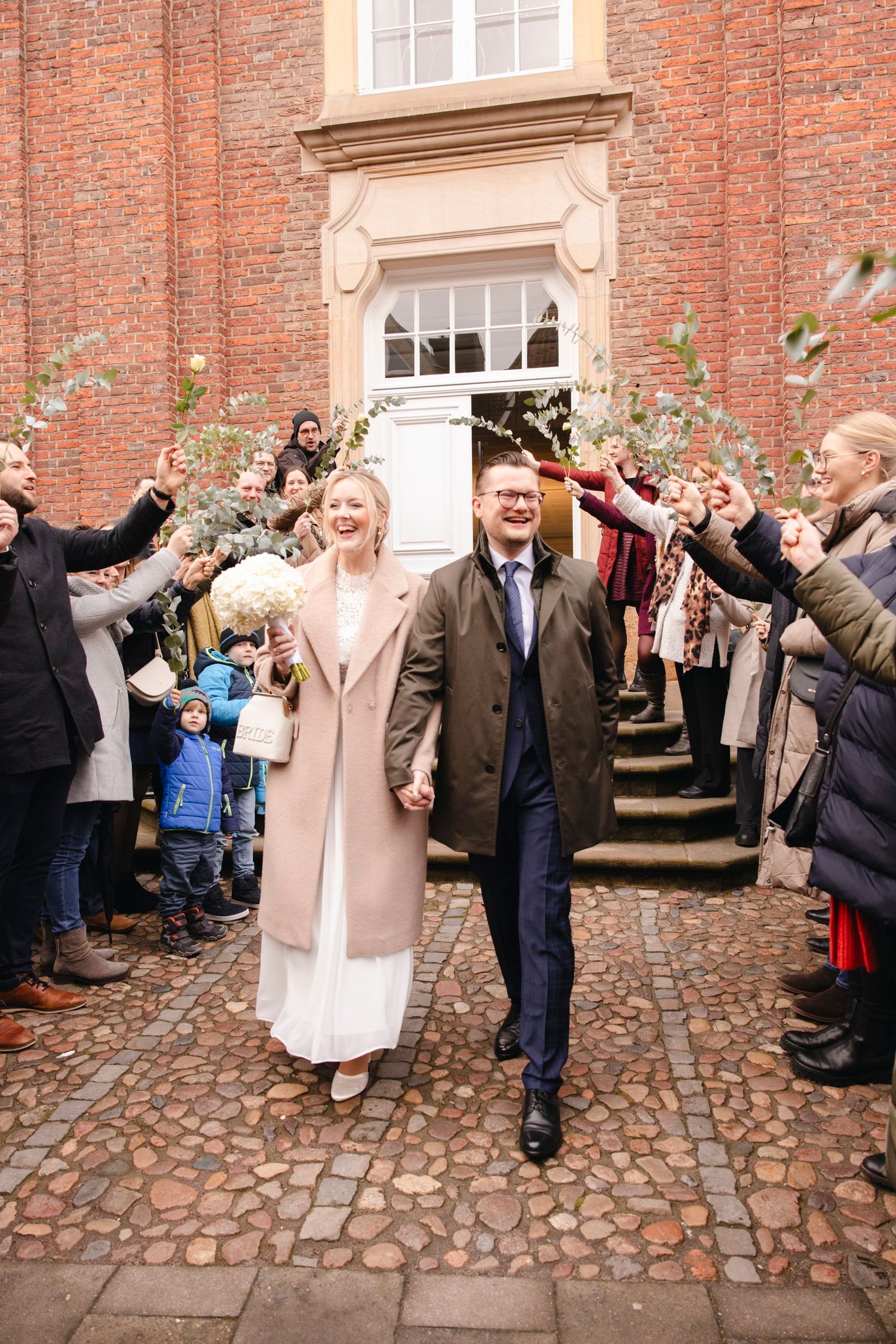 Smiling newlyweds walk hand in hand through cheering guests outside historic brick building.