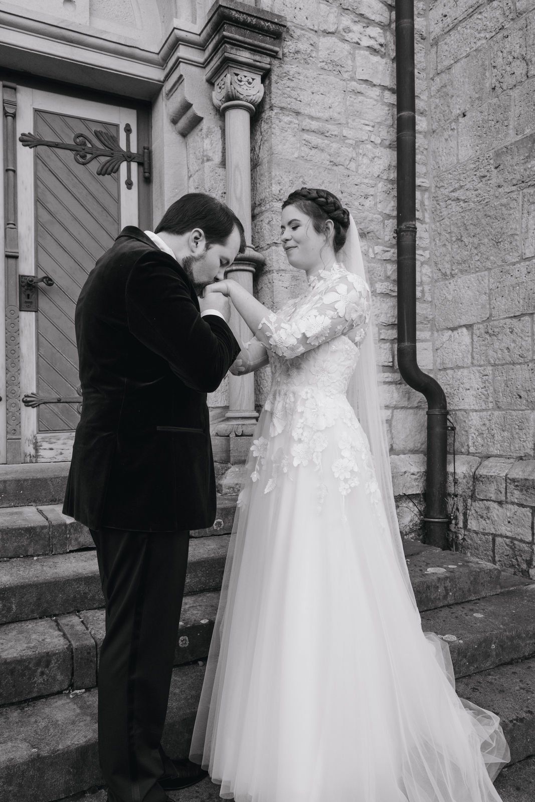 Black and white photo of groom kissing brides hand on church steps in winter wedding portrait.