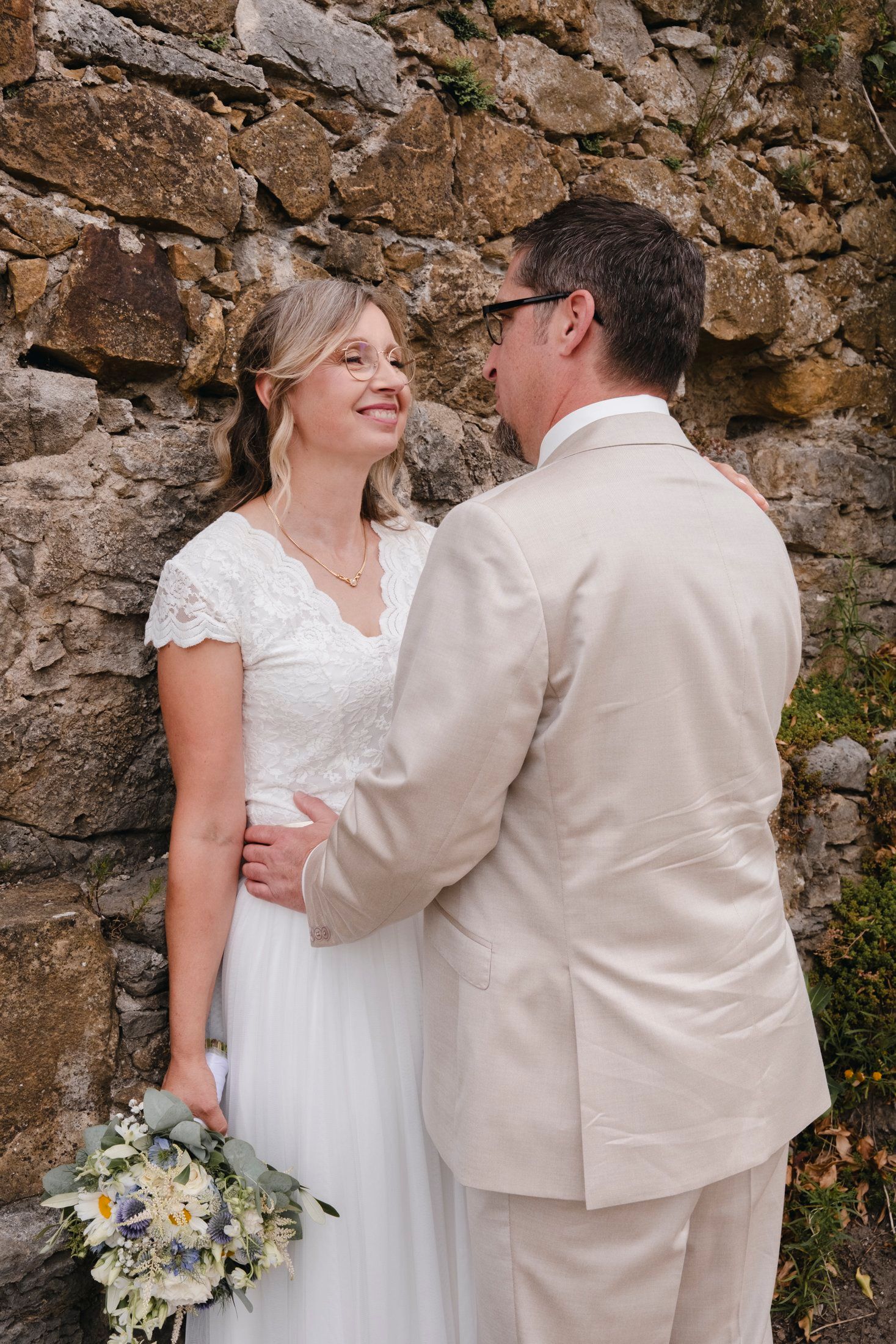 Smiling bride in white lace dress holds bouquet while groom in beige suit embraces her against rustic stone wall.