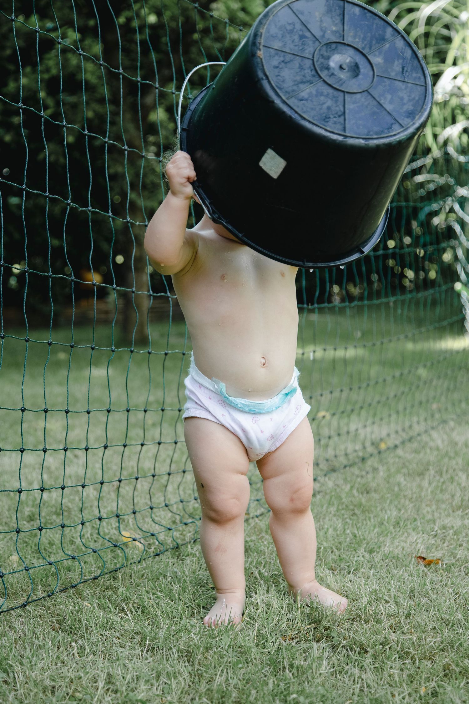 Curious toddler in diaper lifting large black bucket over head while standing on grass outdoors.