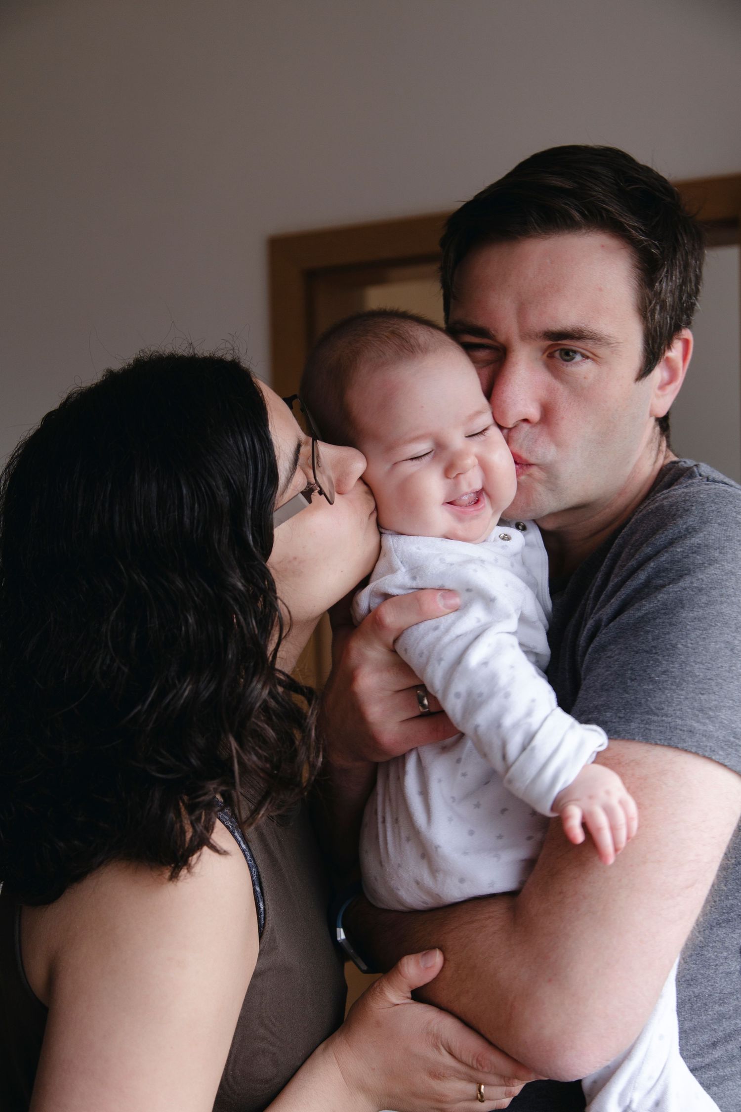 Loving parents kissing their smiling baby on both cheeks simultaneously in cozy home setting.