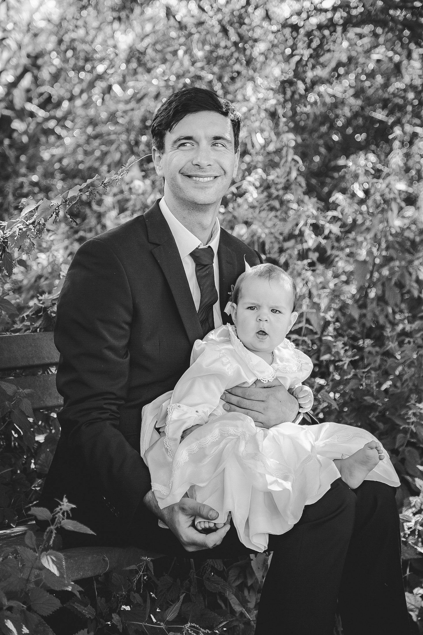Black and white photo of a smiling man in a suit holding a baby in a white christening gown outdoors.