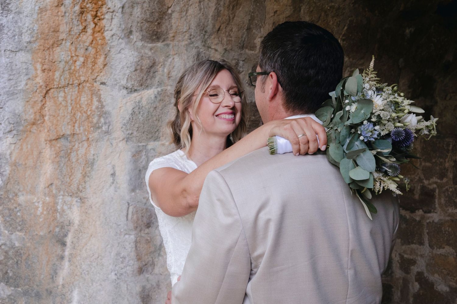 Smiling bride holding a bouquet embraces groom against a rustic stone wall at their wedding.