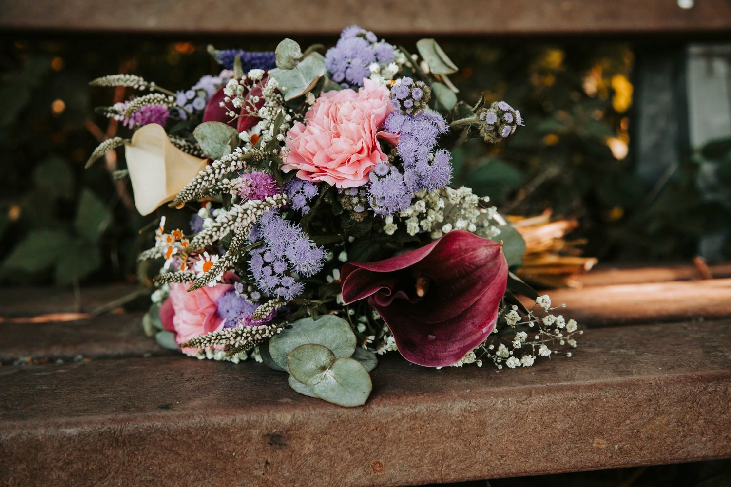 Colorful bridal bouquet with pink roses, purple flowers, and eucalyptus on wooden bench.