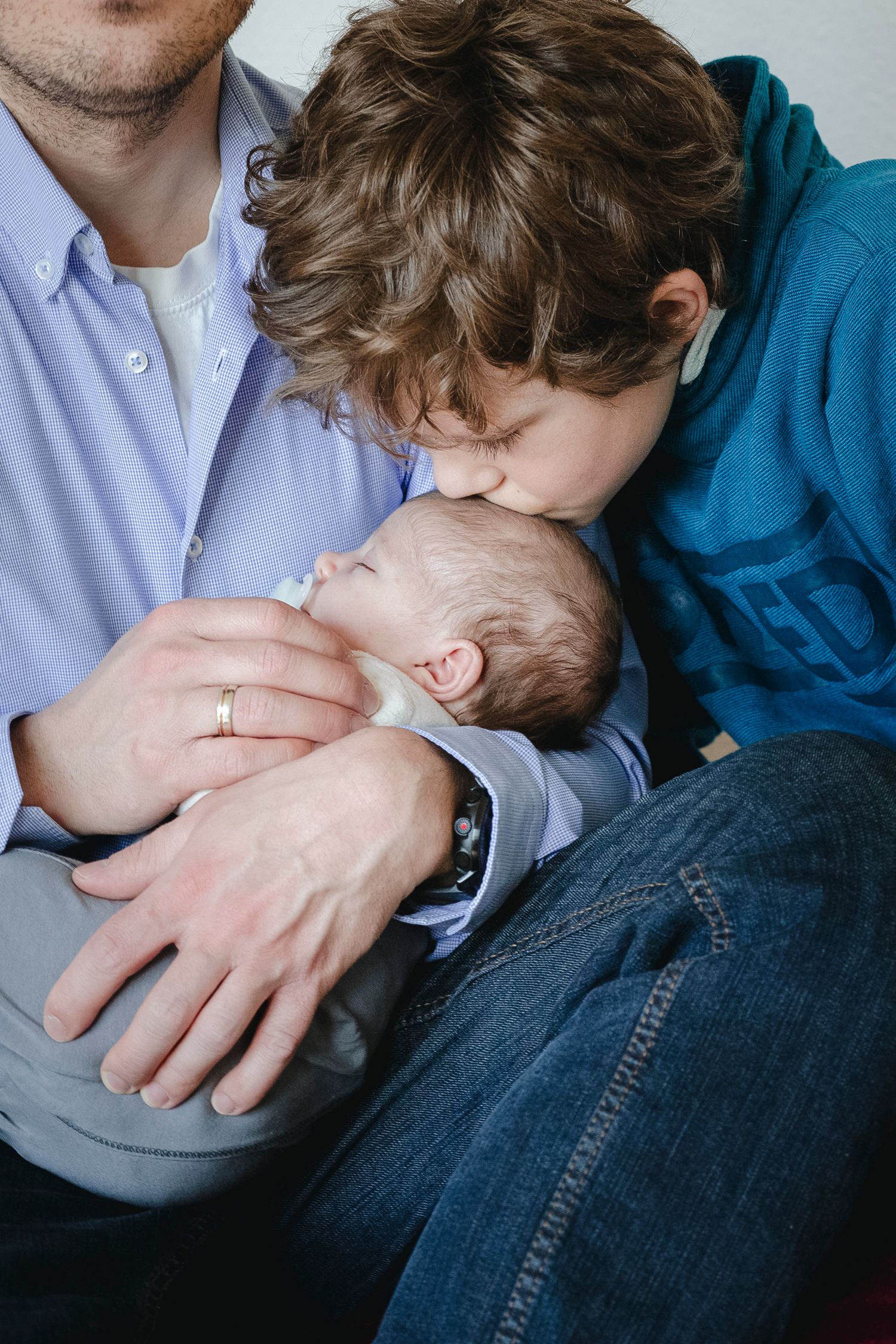 Older brother kissing newborn sibling while father holds baby in his arms.