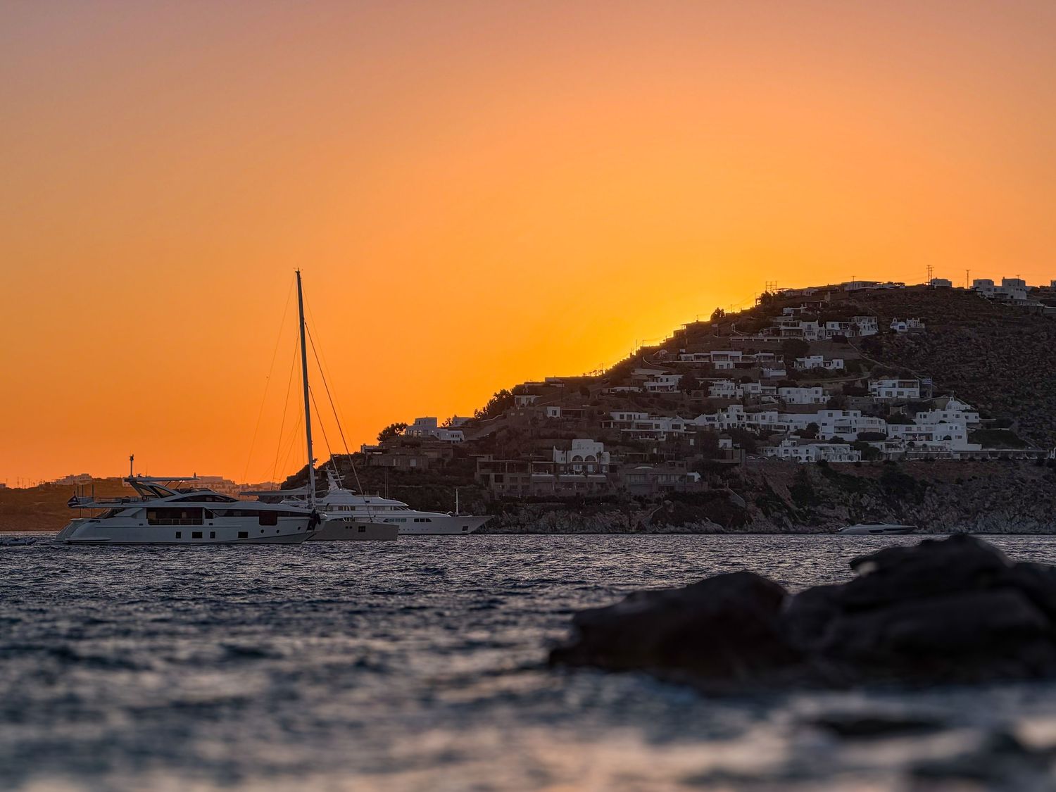 Yacht and sailboat on the Aegean Sea at sunset with whitewashed buildings along the coast of Scorpios Beach in Mykonos, Greece