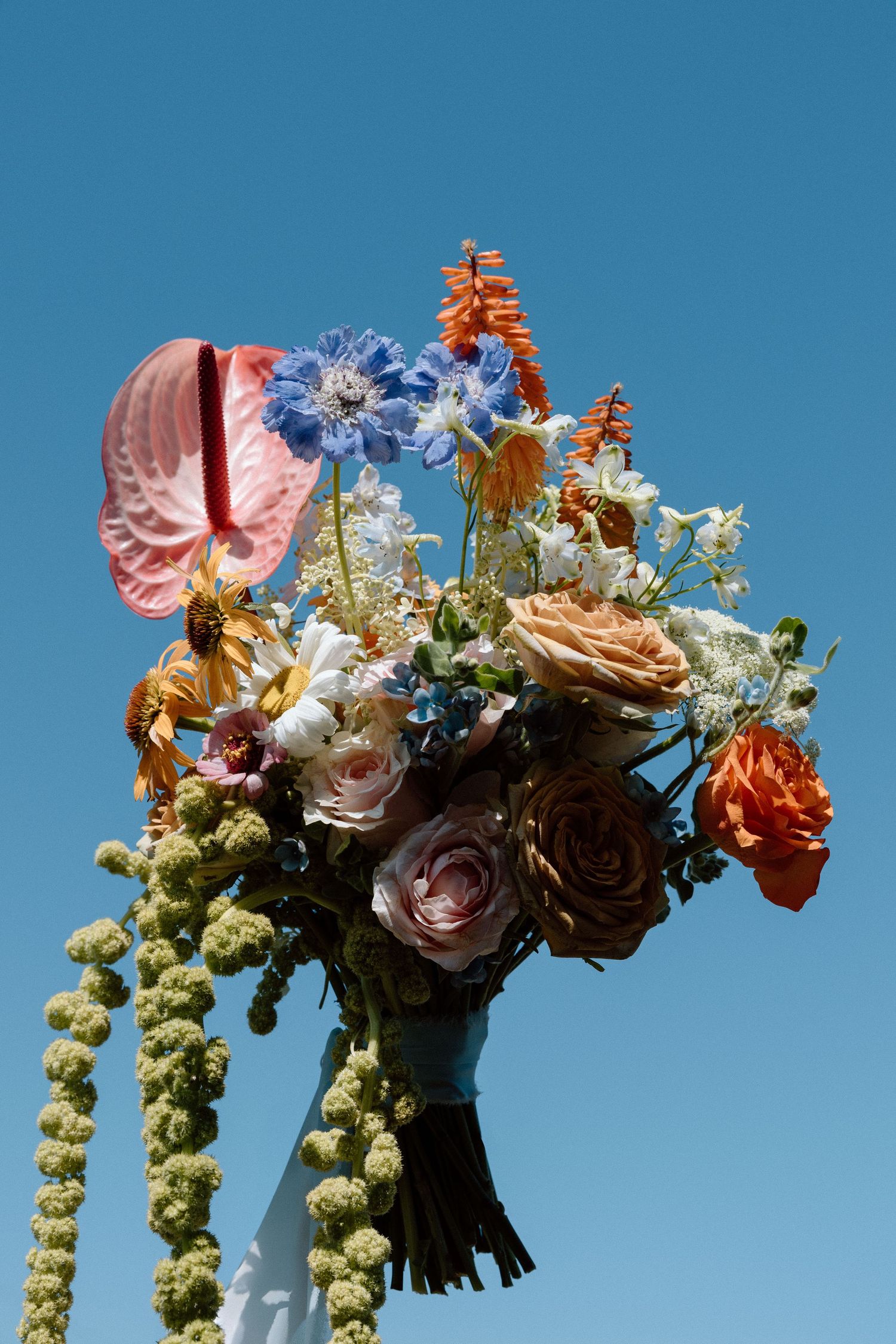 A vibrant floral wedding bouquet with roses, delphiniums and cascading greenery against a bright blue sky background.