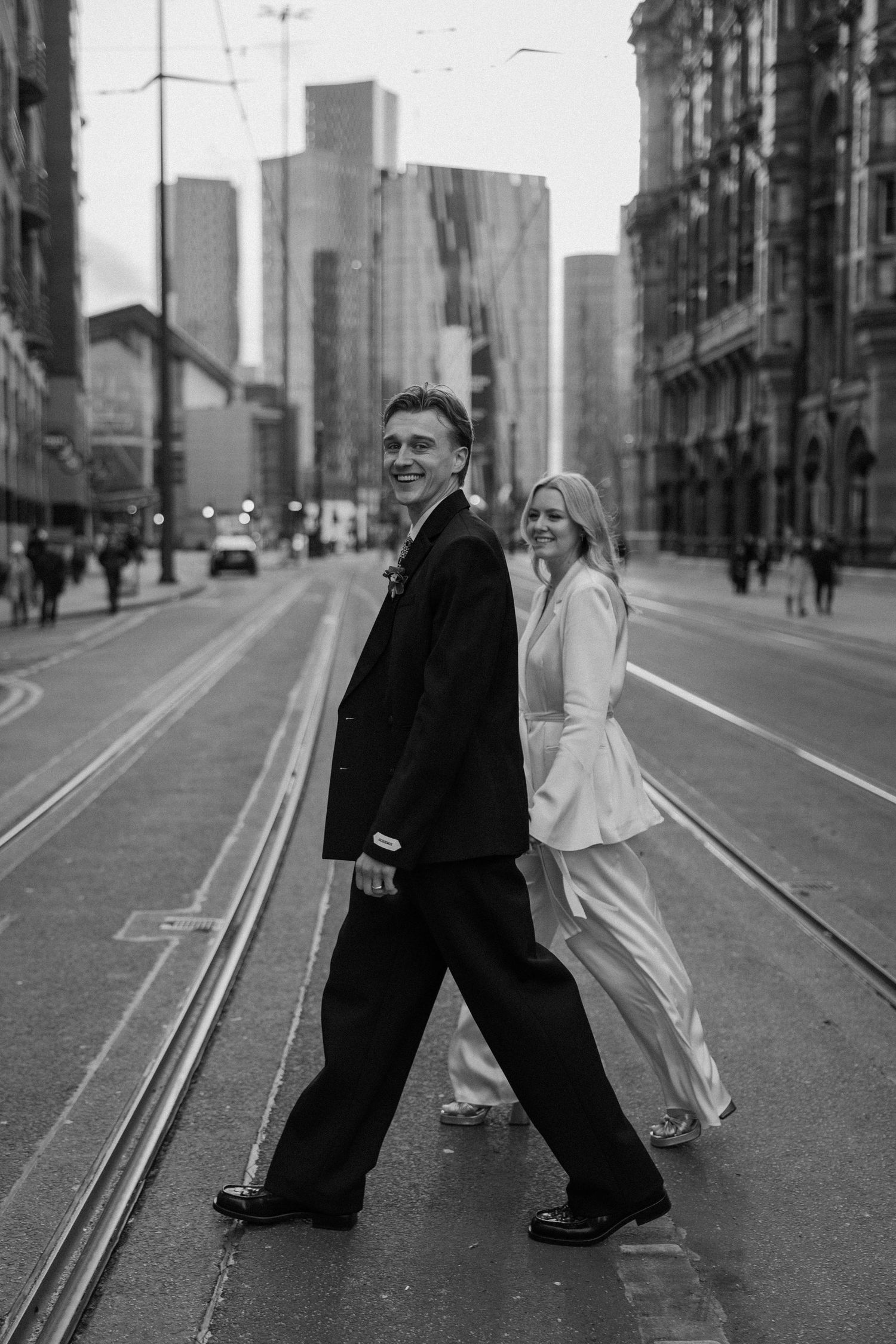 A black and white urban street scene with a couple walking across tram tracks with city buildings in the background at a Manchester wedding.