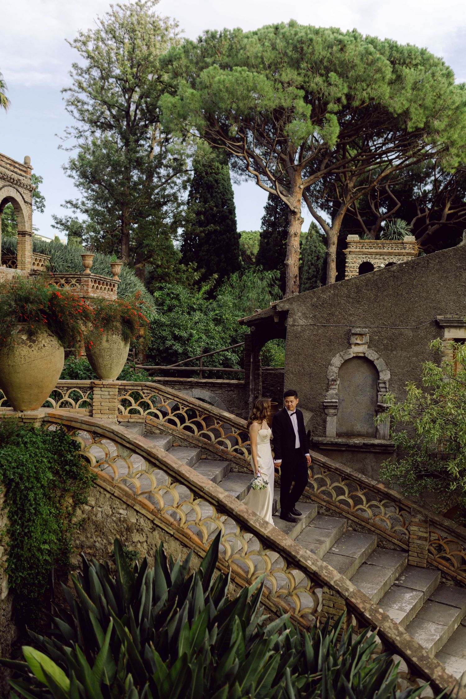 Stone staircase with ornate railing winds through a Mediterranean garden with terracotta pots and pine trees at Taormina Sicily wedding.