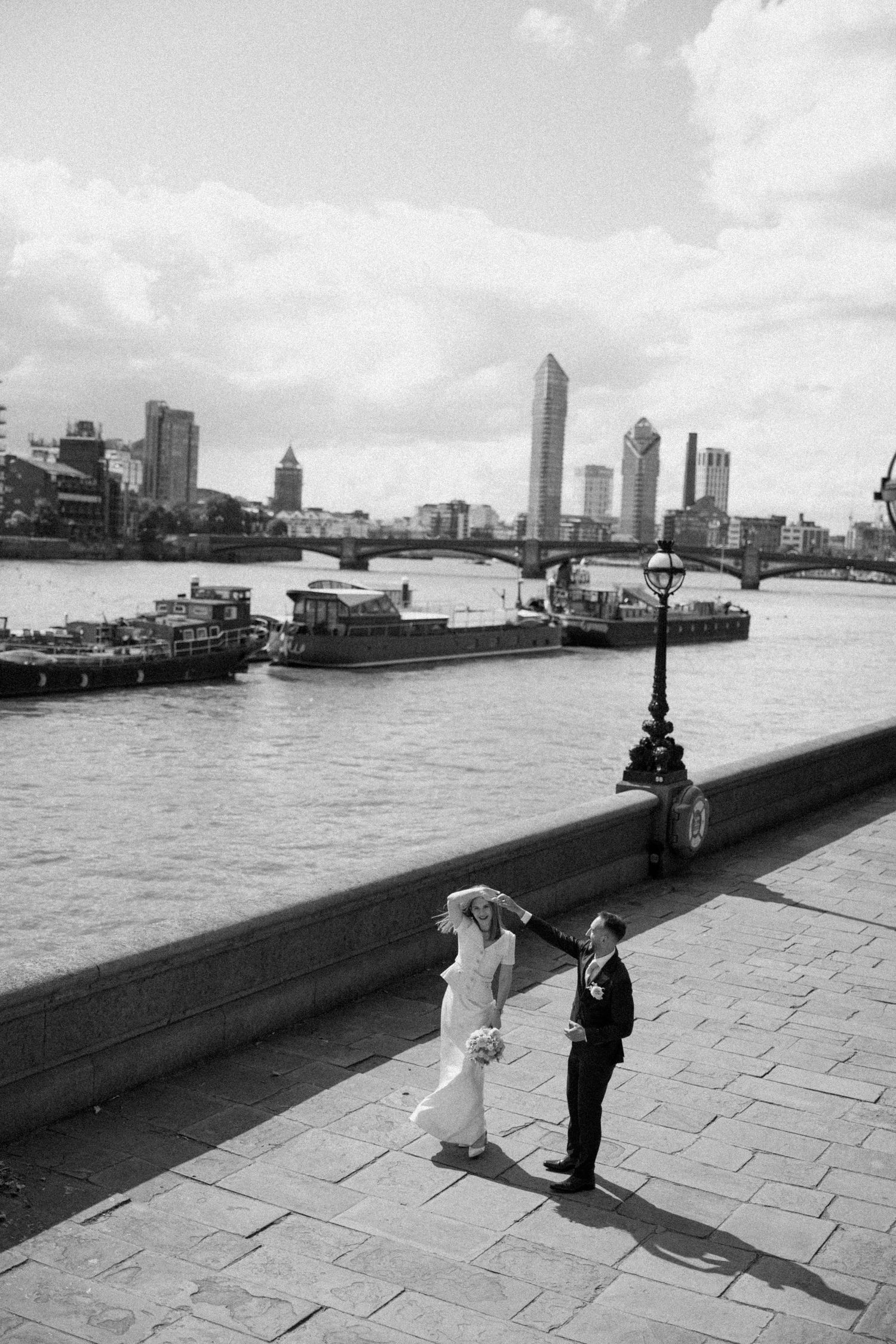Black and white cityscape photo showing two figures on a waterfront promenade with boats and buildings in the background at a London wedding