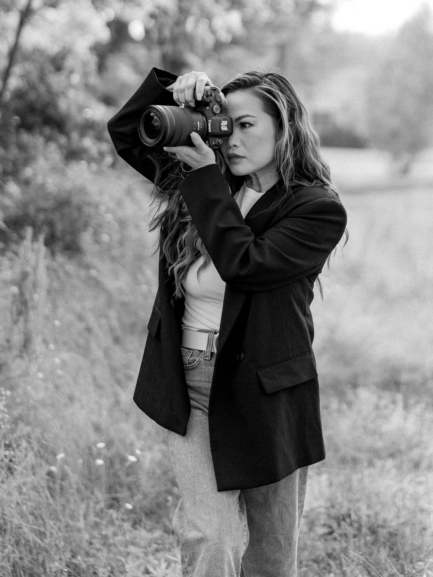 A photographer wearing a black blazer and white top takes pictures with a camera in a natural outdoor setting.
