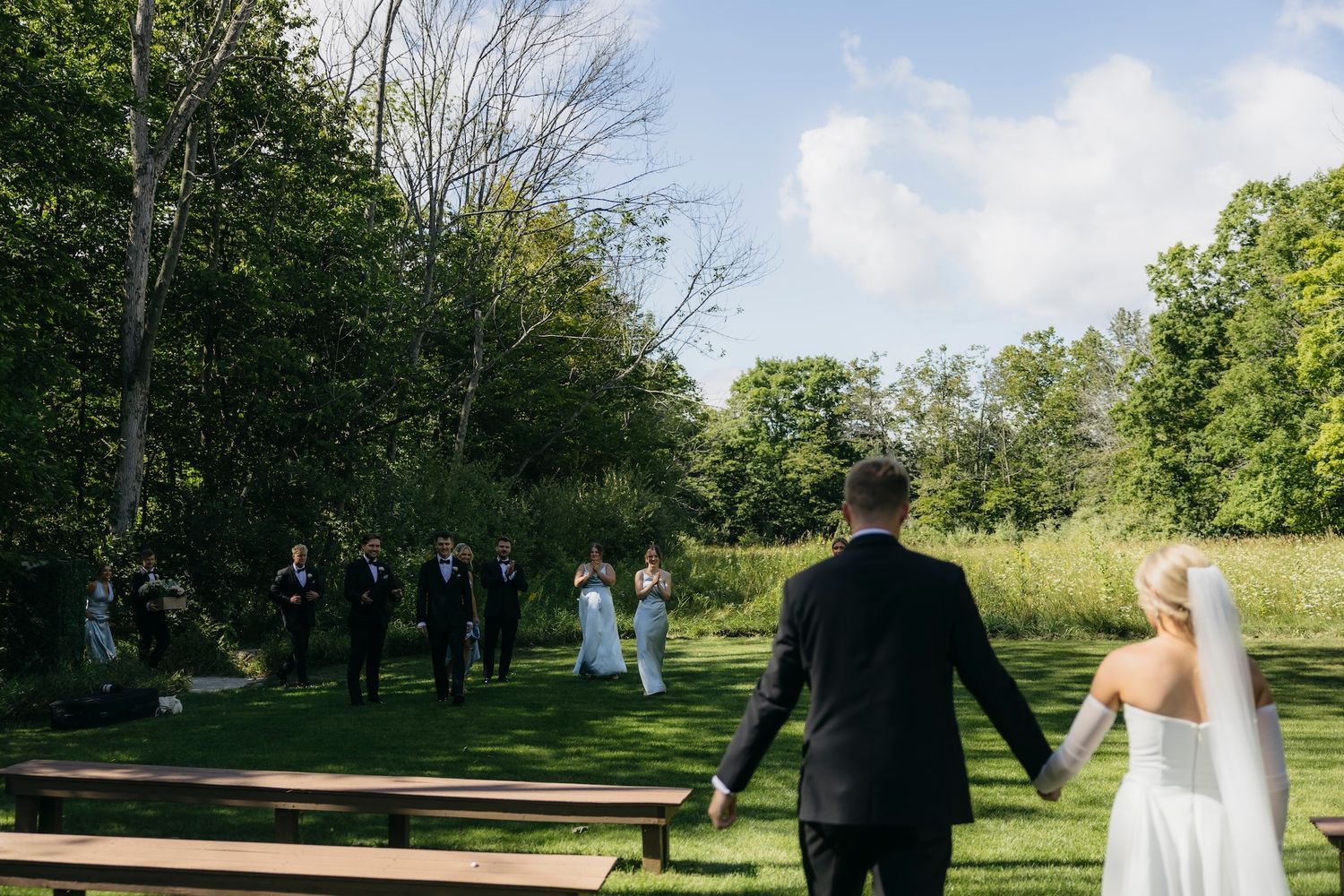 Bride and groom walk hand-in-hand toward wedding guests gathered outdoors in a lush green garden ceremony.