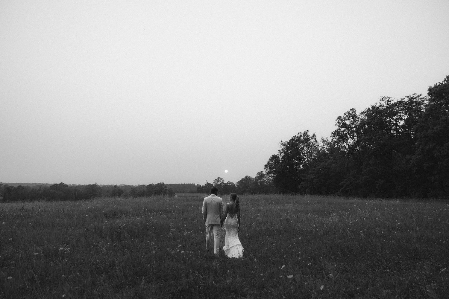 Black and white photo of a couple holding hands walking through a field at dusk with trees in the background.