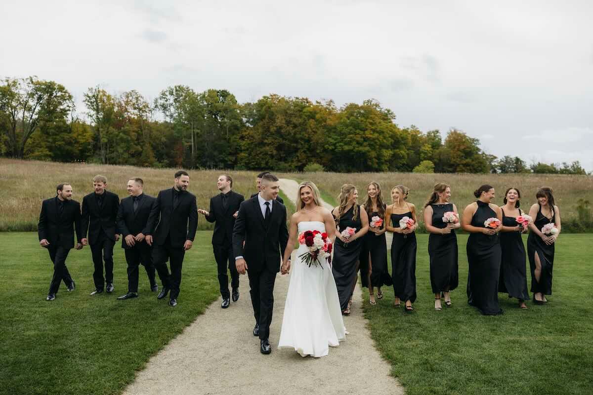 Bride in a white gown holding a bouquet walks with groom, groomsmen in black suits and bridesmaids in black dresses along a grassy path.
