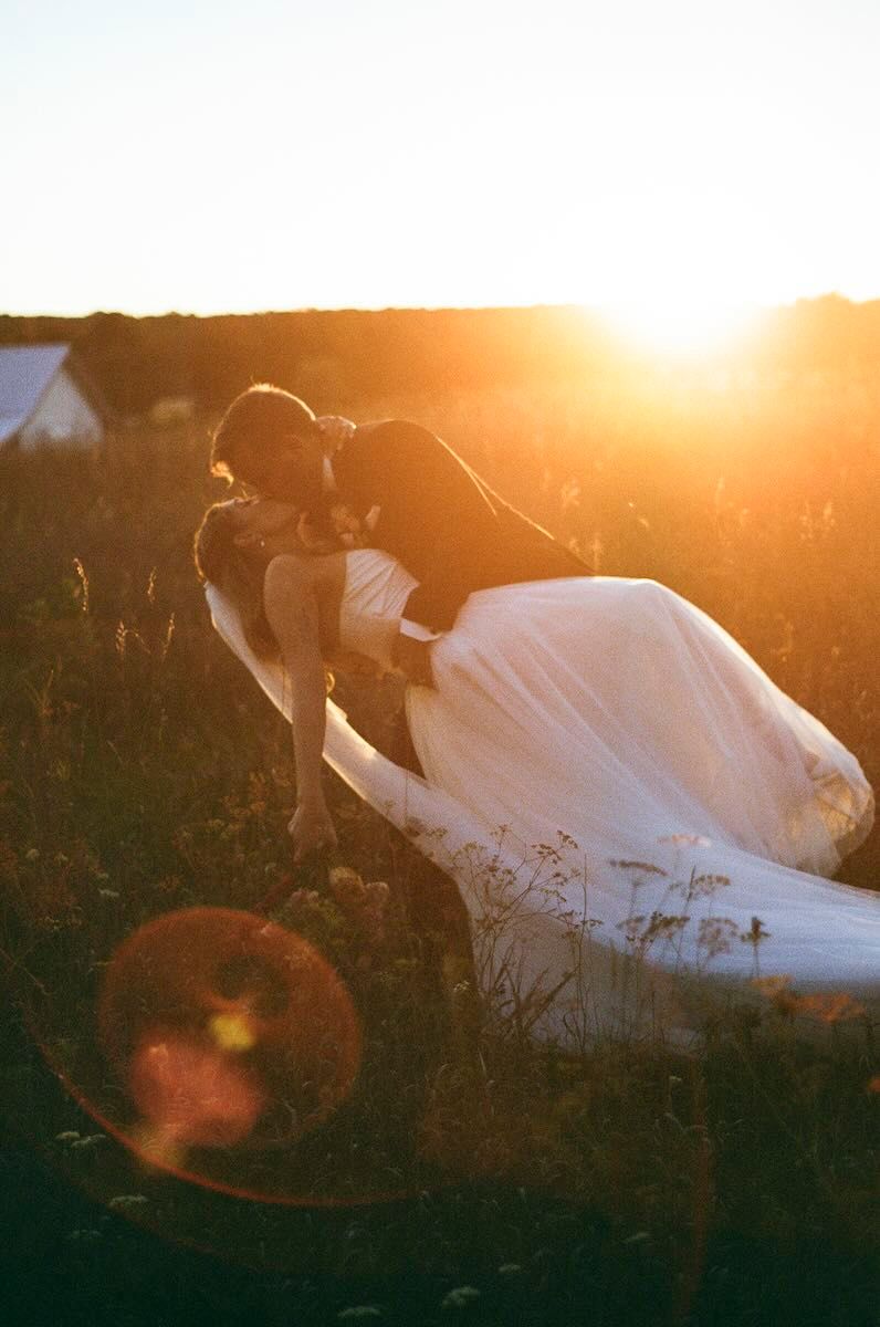 Bride in a flowing white tulle gown shares a romantic dipped kiss in a golden meadow at sunset with warm sun flare.