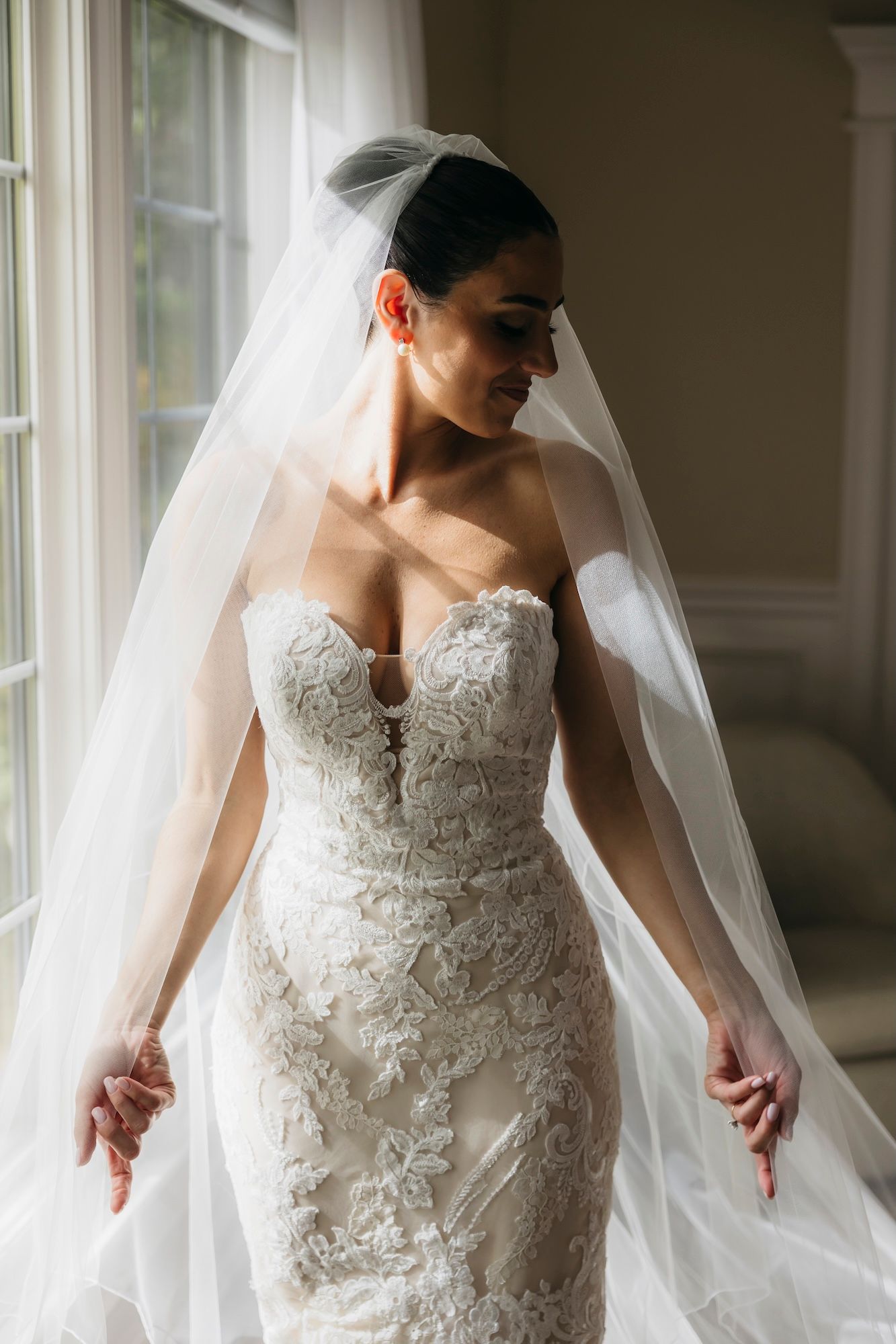 Bride in strapless lace wedding gown and flowing veil poses by window in soft natural light.