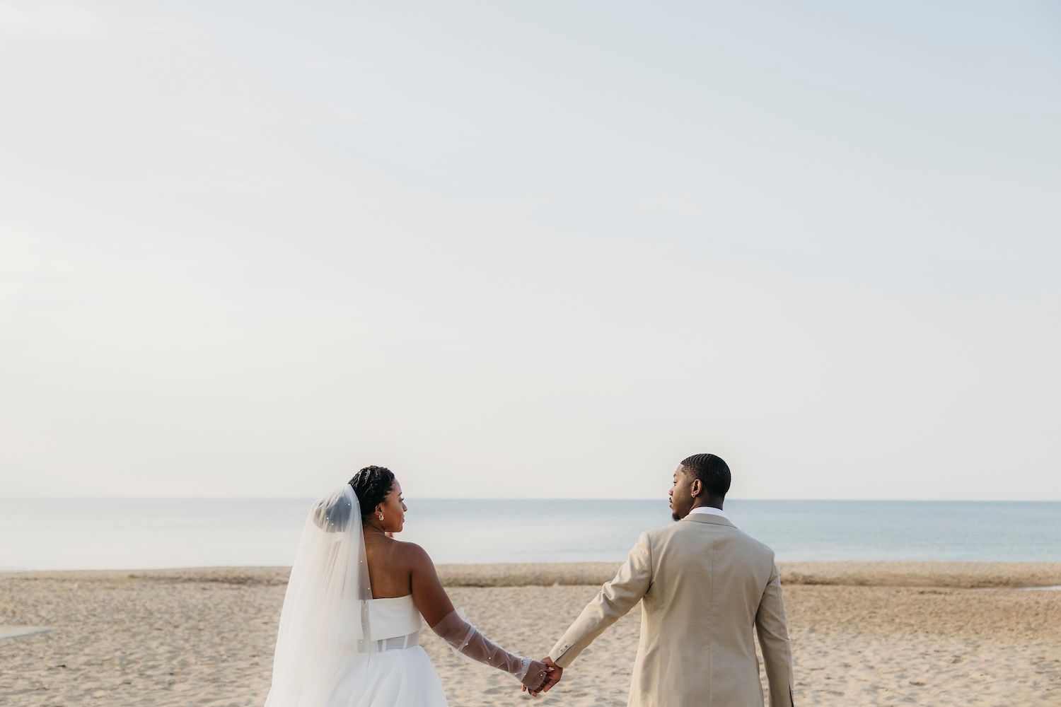 A bride and groom hold hands facing the ocean on a sandy beach, wearing a white gown and tan suit.