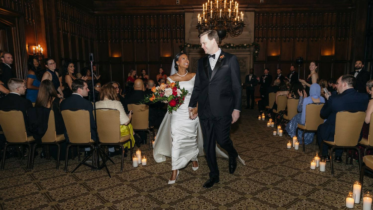 Bride and groom walk down the aisle at elegant candlelit wedding reception in grand ballroom.