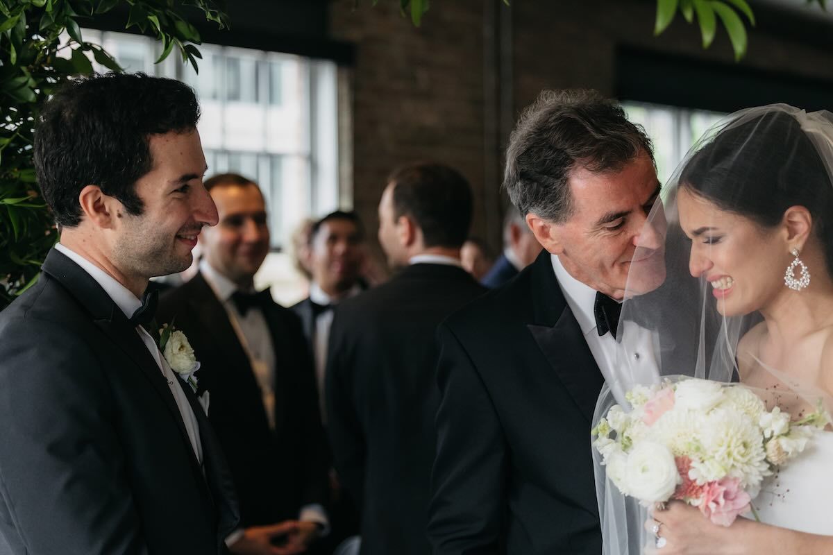 Smiling bride in a veil holding a pink and white bouquet greets a groom in a black tuxedo while wedding guests mingle behind them.