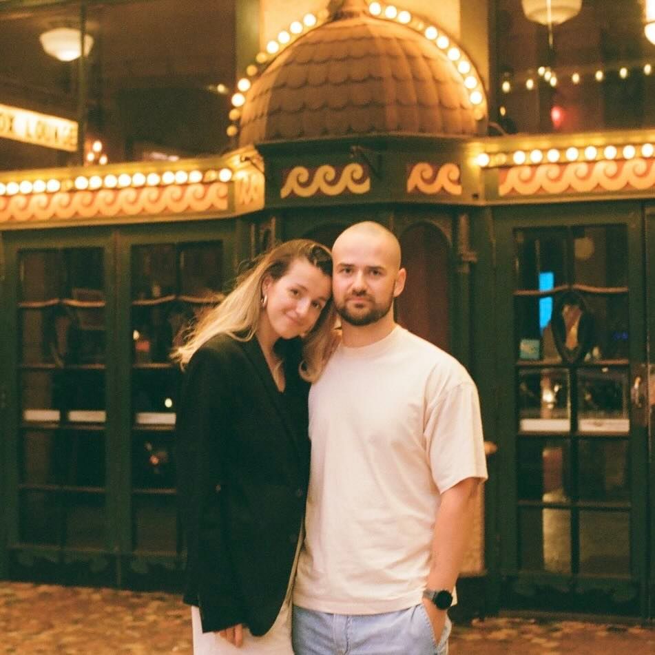 A smiling couple poses close together in front of an ornate vintage building with a domed roof, glowing marquee lights, and glass doors.