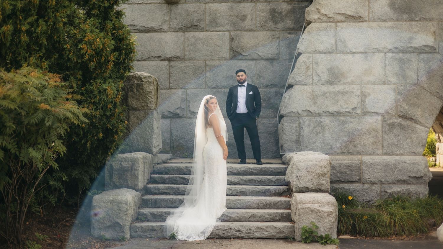 Bride in white gown and veil stands on stone steps with groom in tuxedo behind her at outdoor wedding venue.