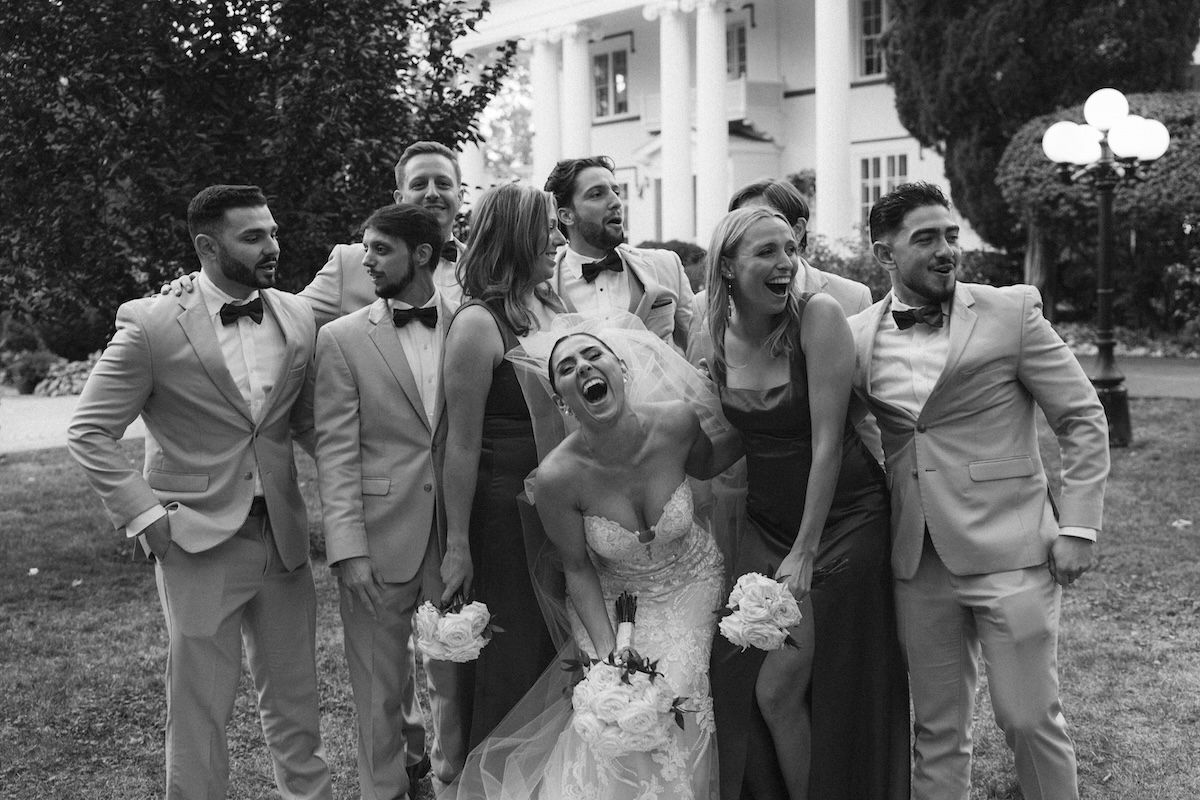 Black and white wedding party photo with bride, bridesmaids, and groomsmen laughing joyfully outside elegant venue.