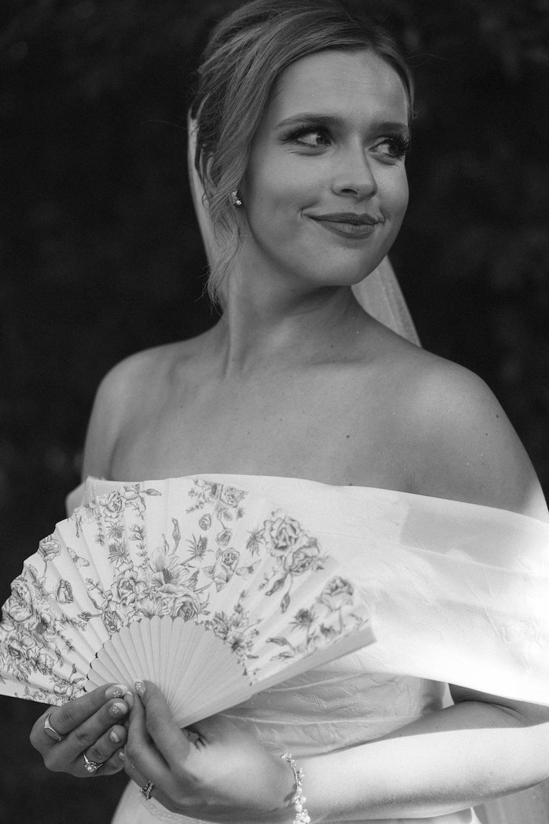Black and white portrait of a bride holding a floral hand fan, wearing an off-shoulder gown and veil.