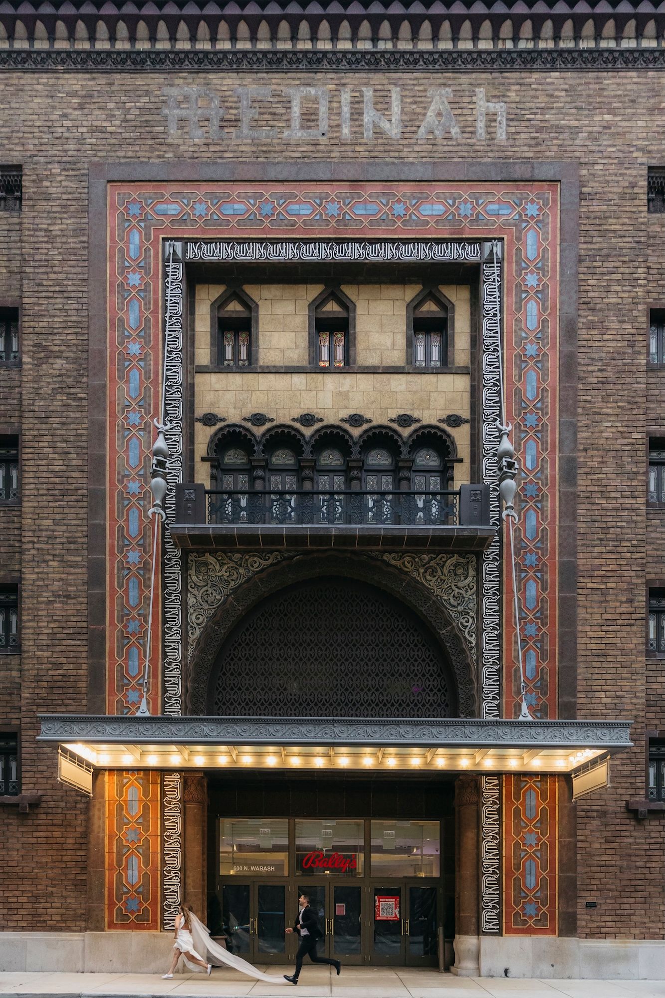 Bride in flowing white gown walks past ornate Medinah Temple entrance with decorative Islamic-inspired brick facade in Chicago.