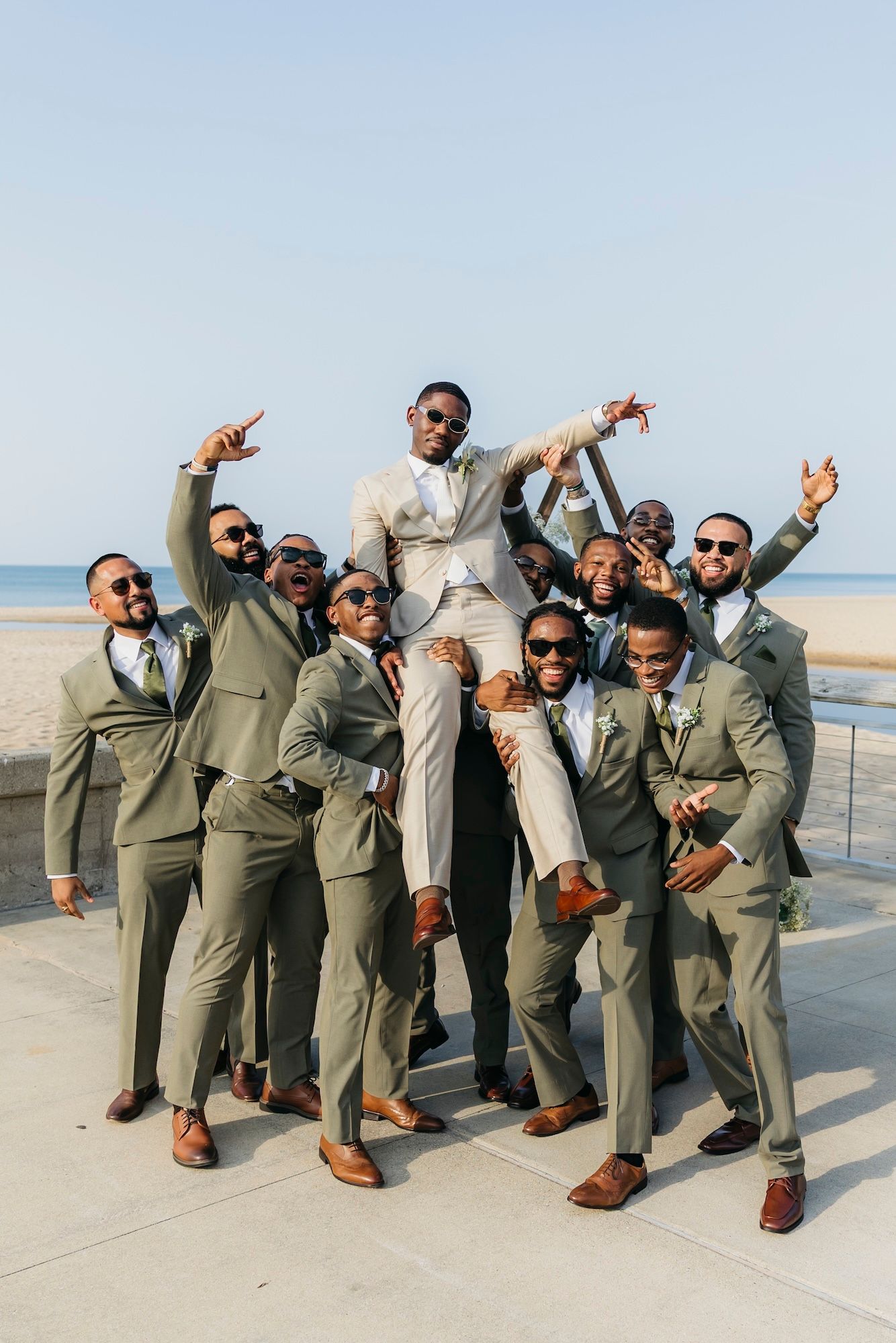 Groom lifted by groomsmen in olive suits near beach, celebrating with raised hands and sunglasses on sunny day.