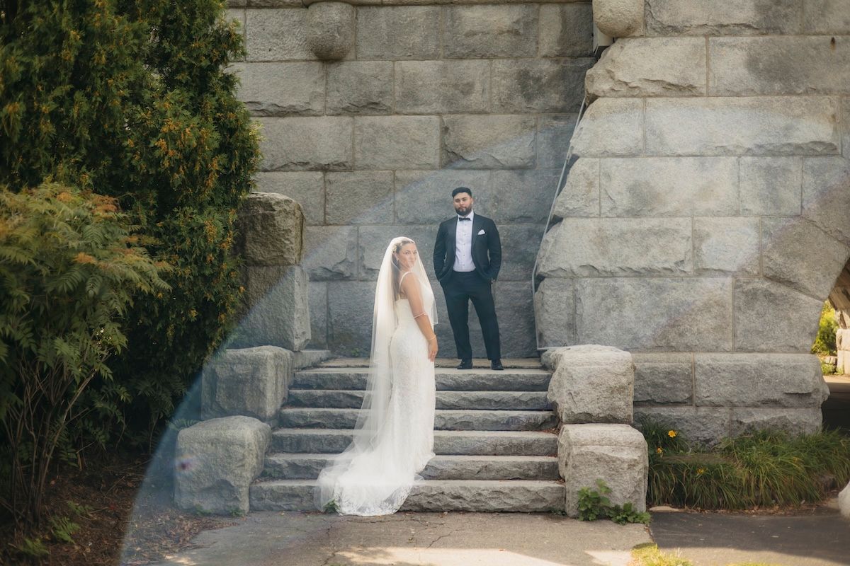 Bride in white gown and veil stands on stone steps with groom in navy suit at historic outdoor wedding venue.