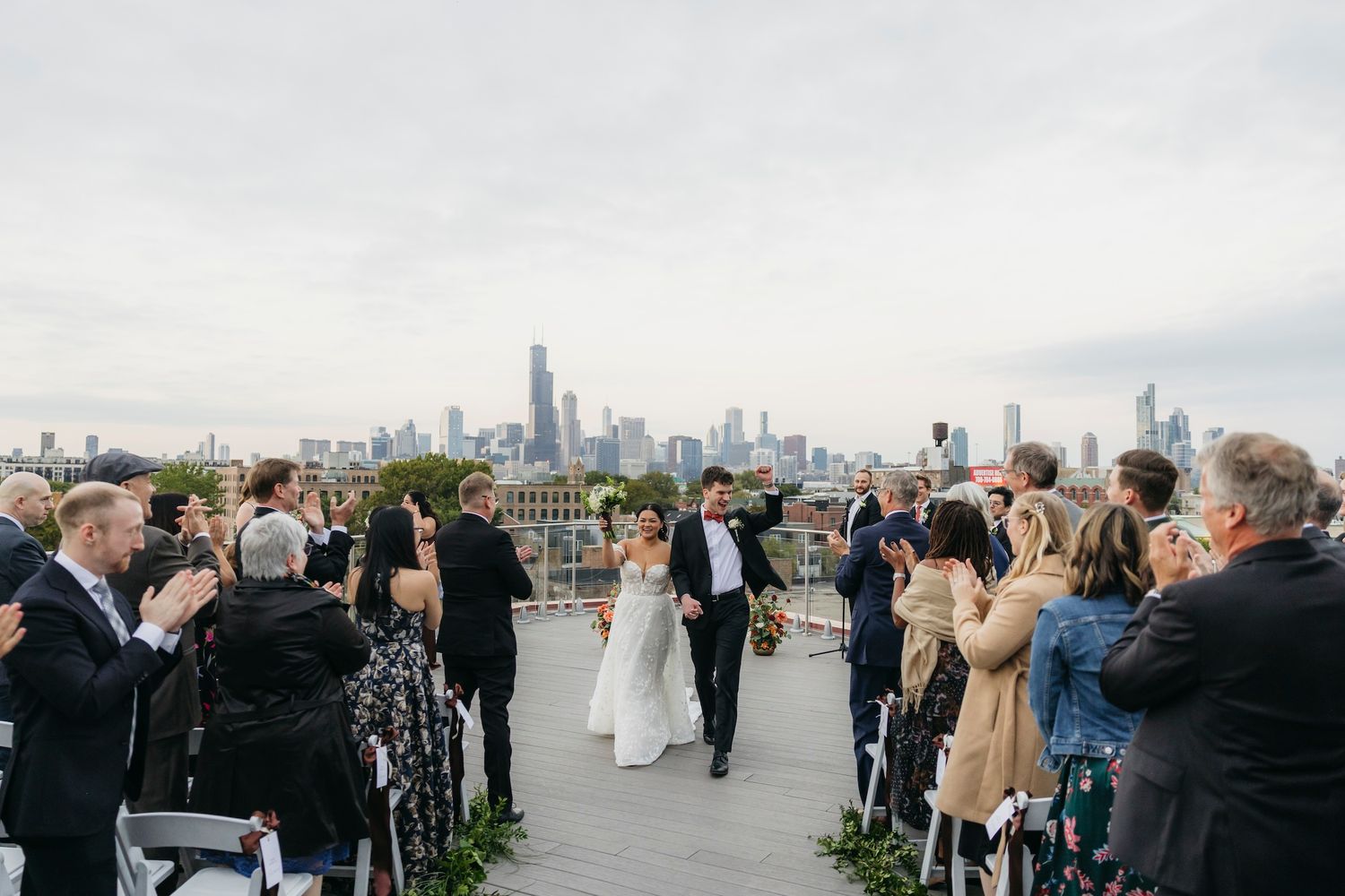 Couple walks down aisle at rooftop wedding ceremony with Chicago skyline backdrop, guests applauding in celebration.