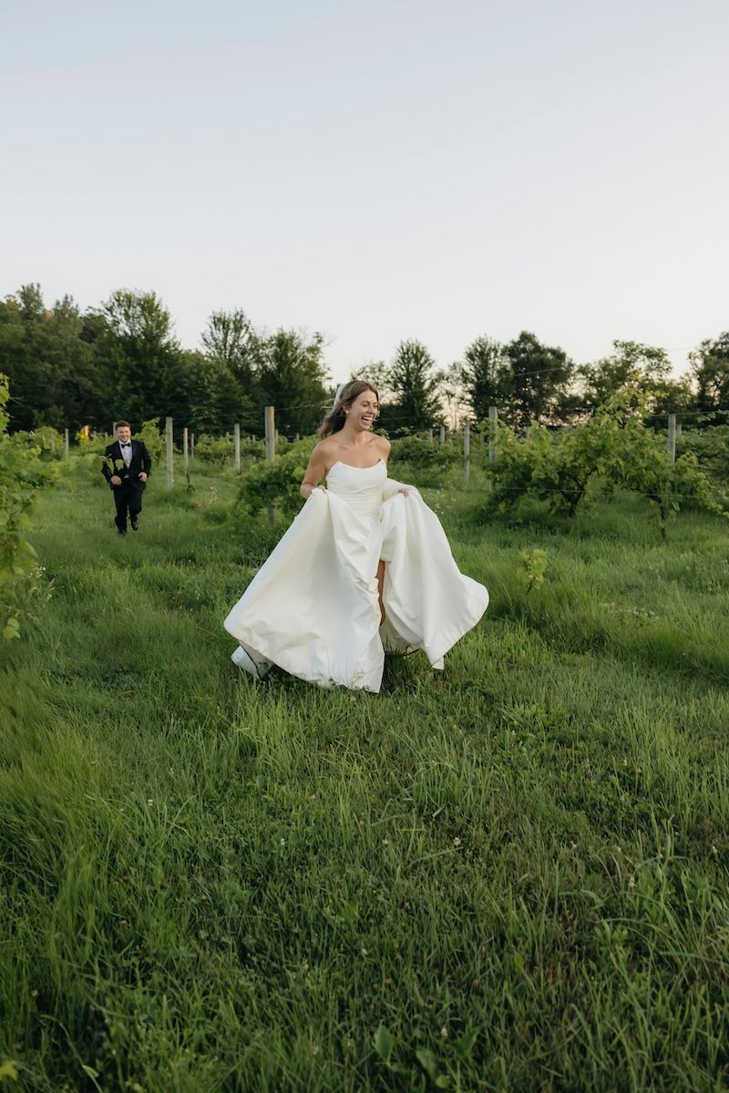 Bride in white gown runs through lush vineyard rows as groom in tuxedo follows during outdoor wedding.