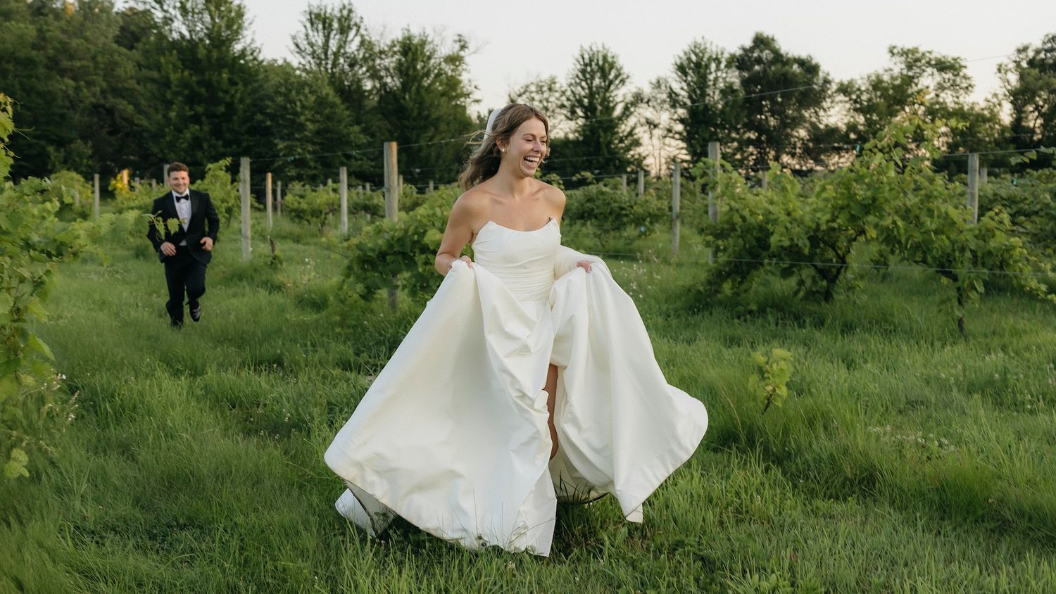 Bride in white gown runs through lush vineyard rows as groom in tuxedo follows during outdoor wedding.